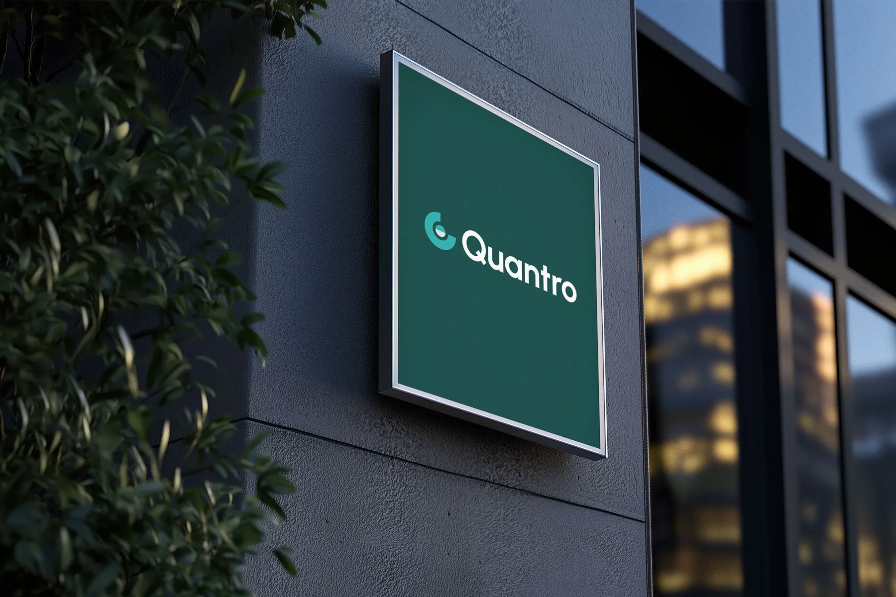 Quantro logo sign mounted on a dark office building wall beside green leaves.
