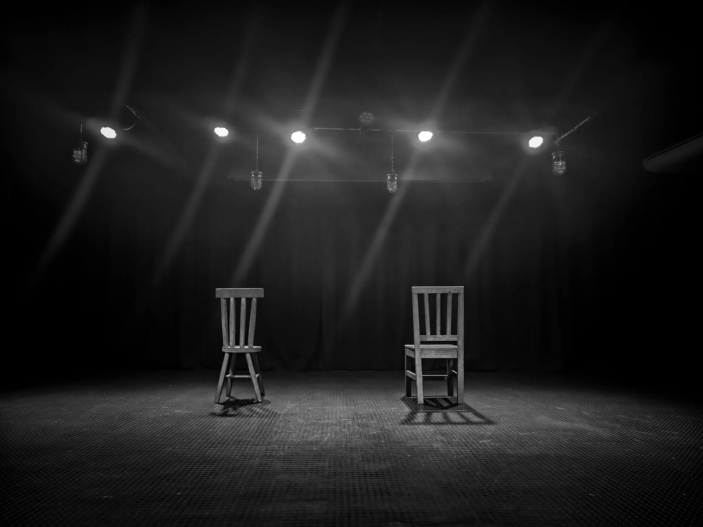 Two wooden chairs on an empty stage under a row of spotlights, in black and white.