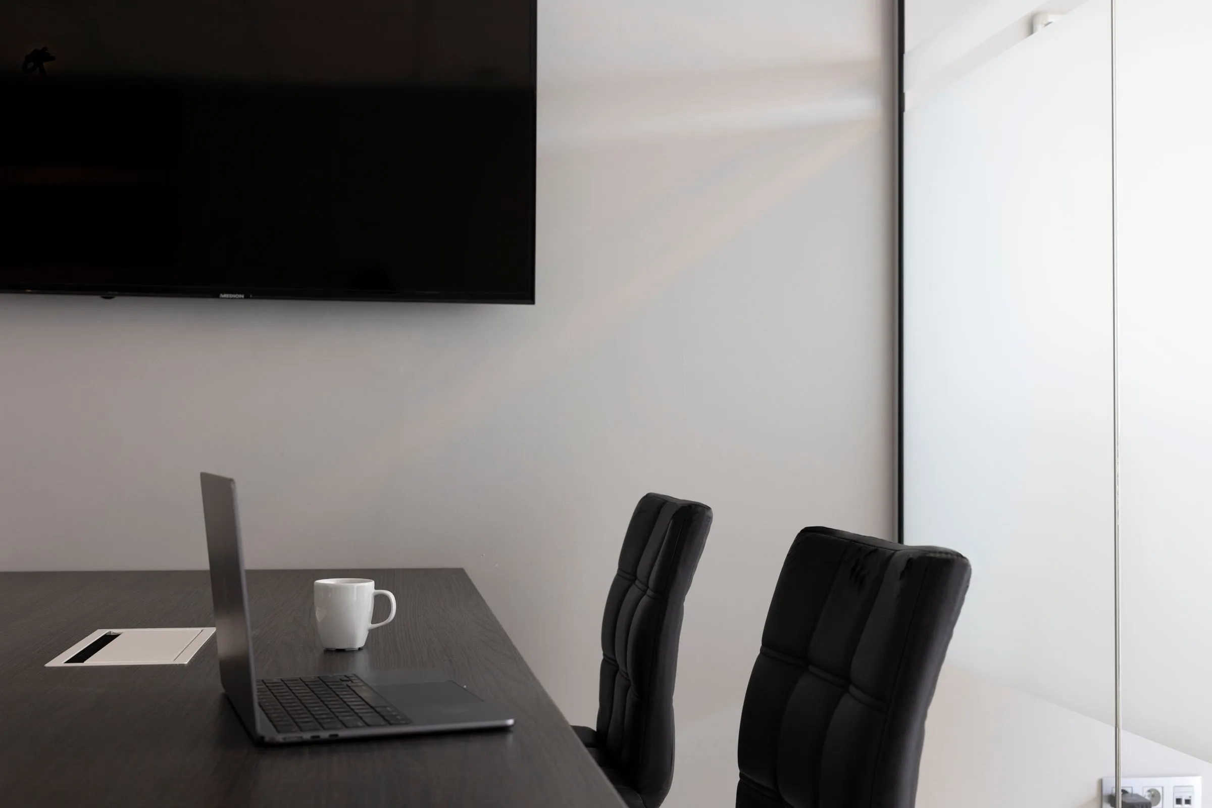 Laptop and white mug on a dark meeting table with black chairs and a wall-mounted screen.
