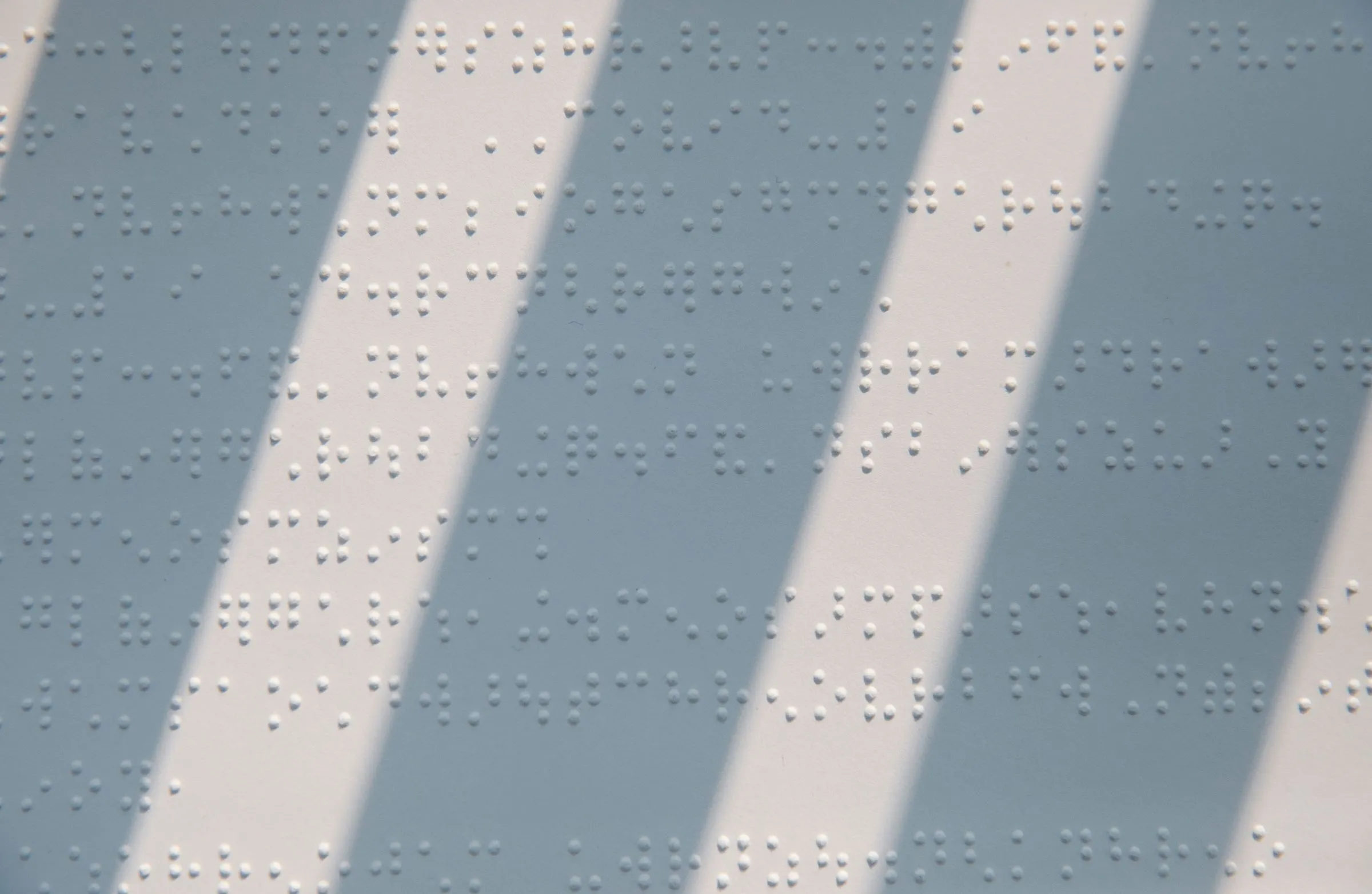 Page of braille text crossed by diagonal stripes of light and blue-grey shadow.