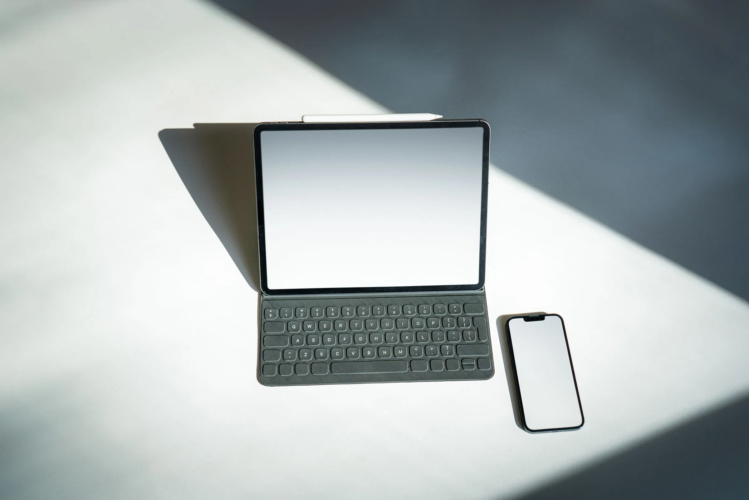 Tablet with a keyboard case and a smartphone on a white table in strong diagonal sunlight.