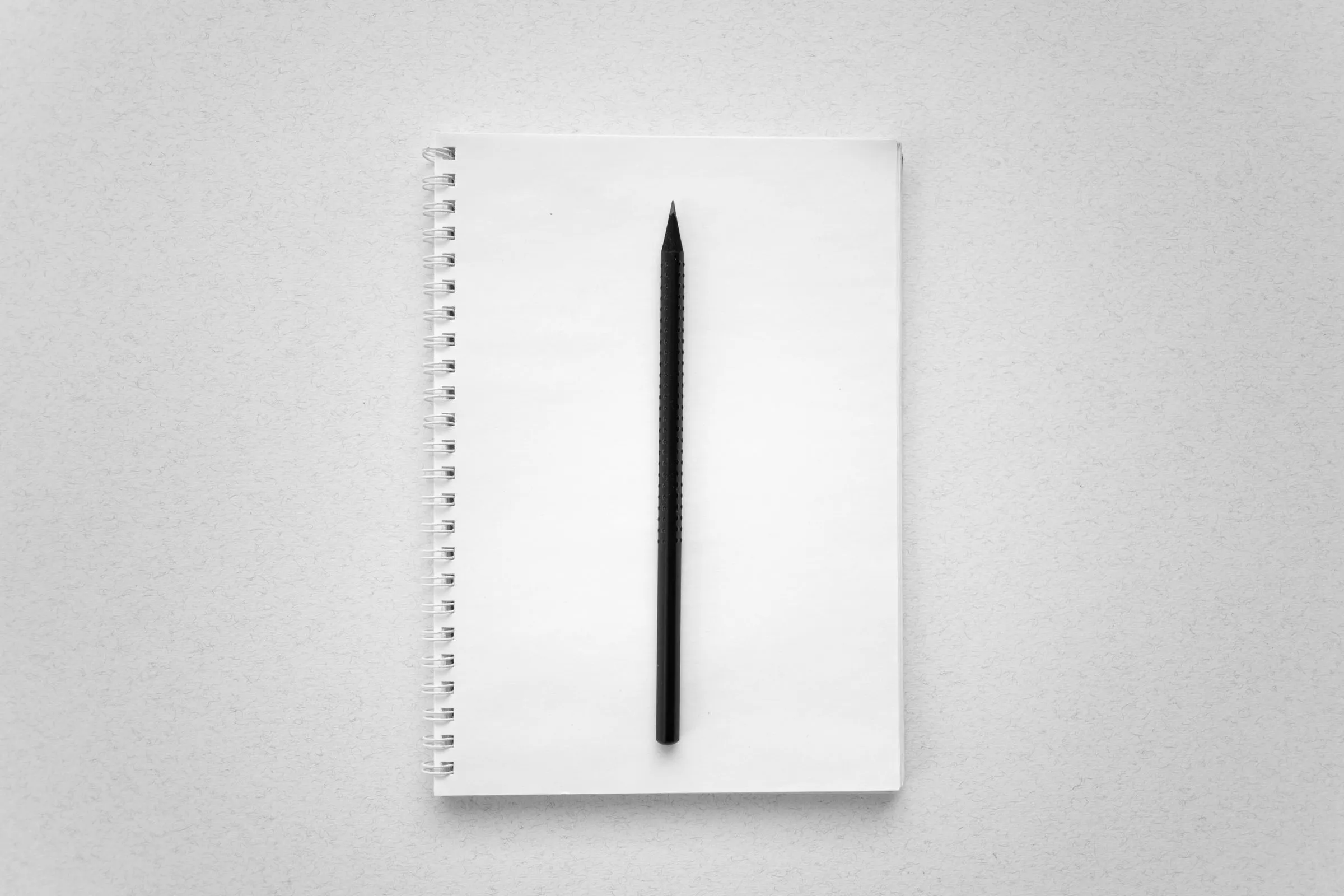 Single black pencil lying on a blank spiral notebook on a white surface.