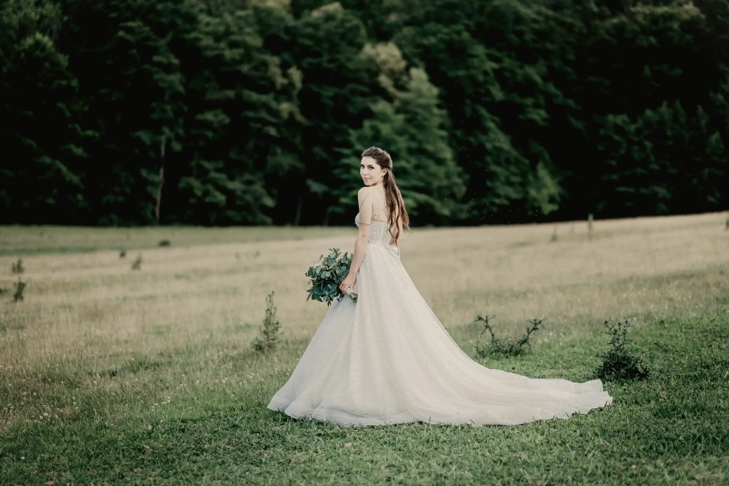Bride in a full off-the-shoulder ball gown stands alone in a green summer meadow, a dark forest edge behind her.