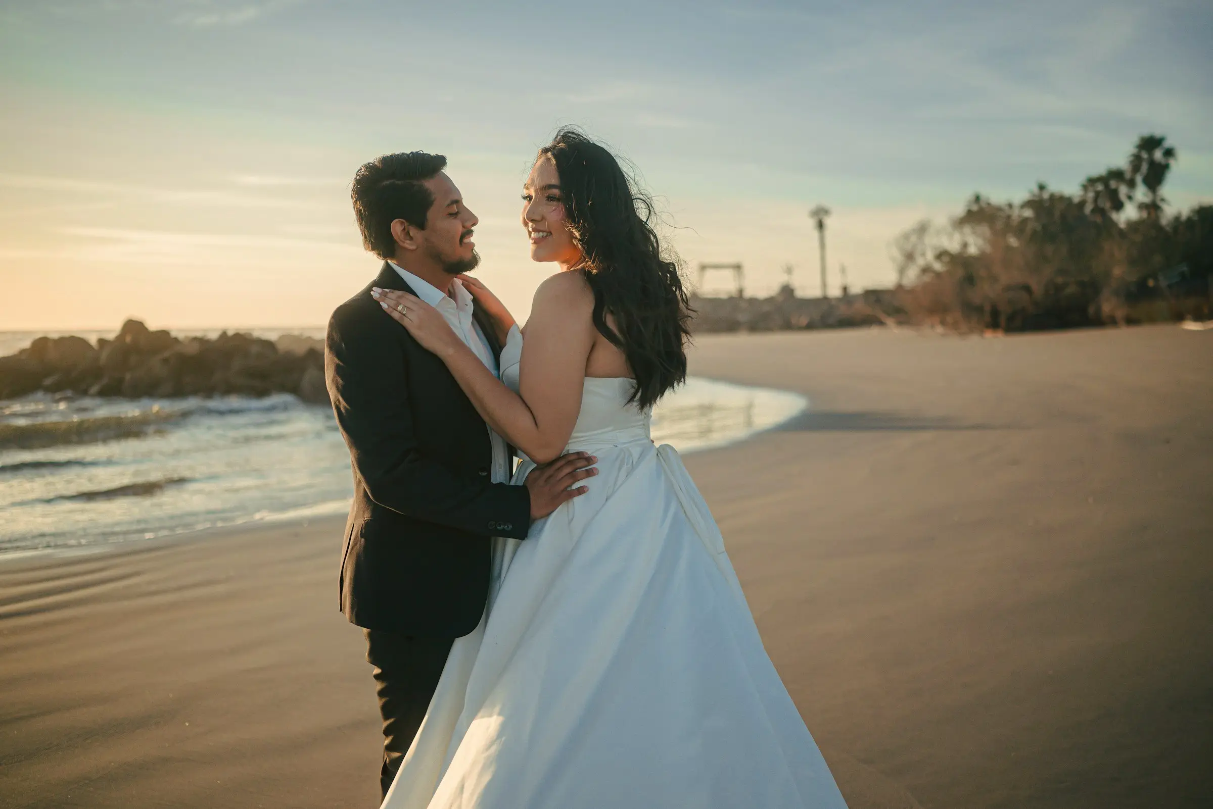 Bride in a strapless satin gown and groom in a dark suit embrace on a wide sandy beach at sunset, palms and low rocks behind them, warm side light.