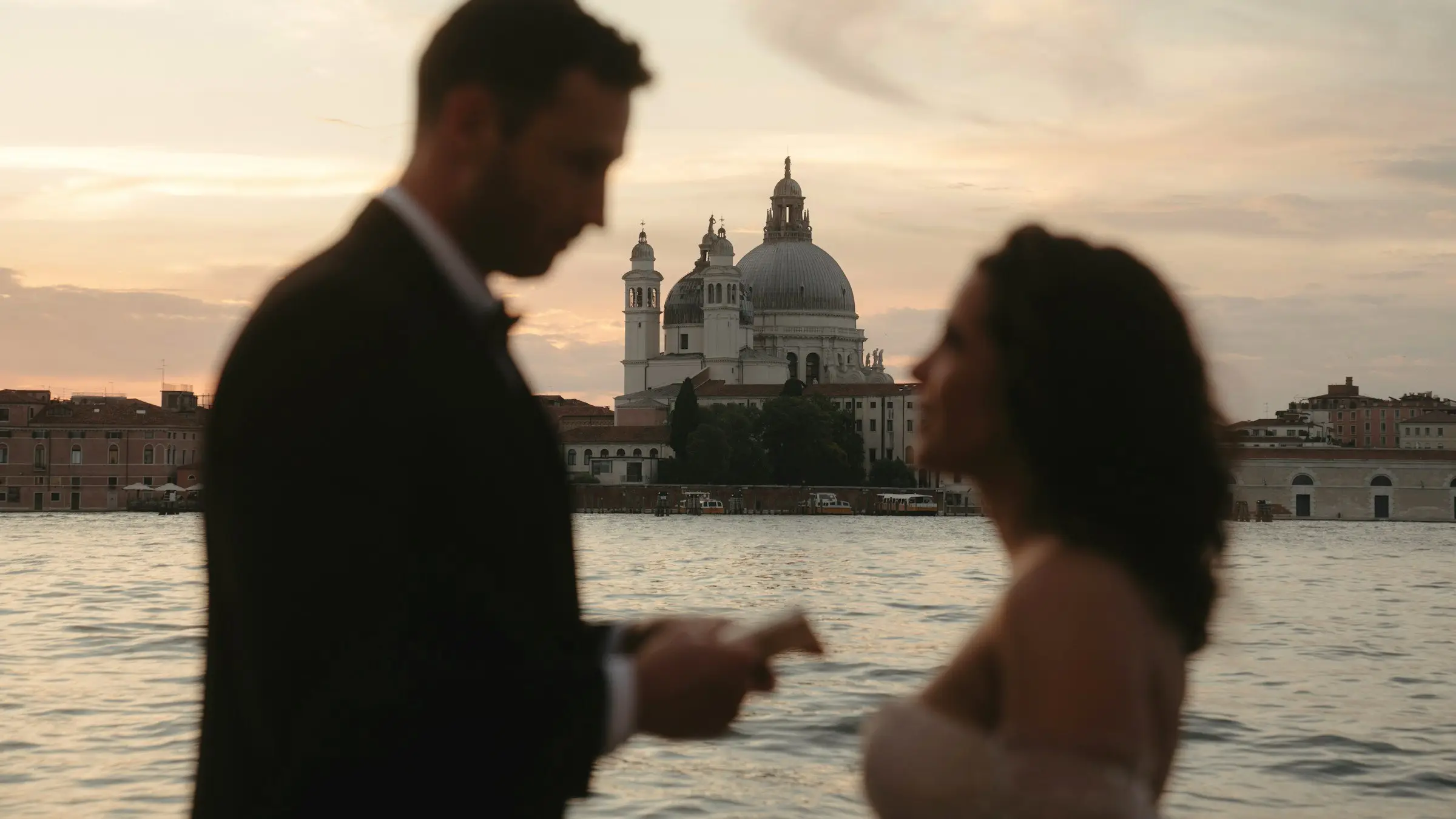 Groom reads his vows to the bride in silhouette on the Venetian waterfront, the domed Santa Maria della Salute glowing at sunset behind.