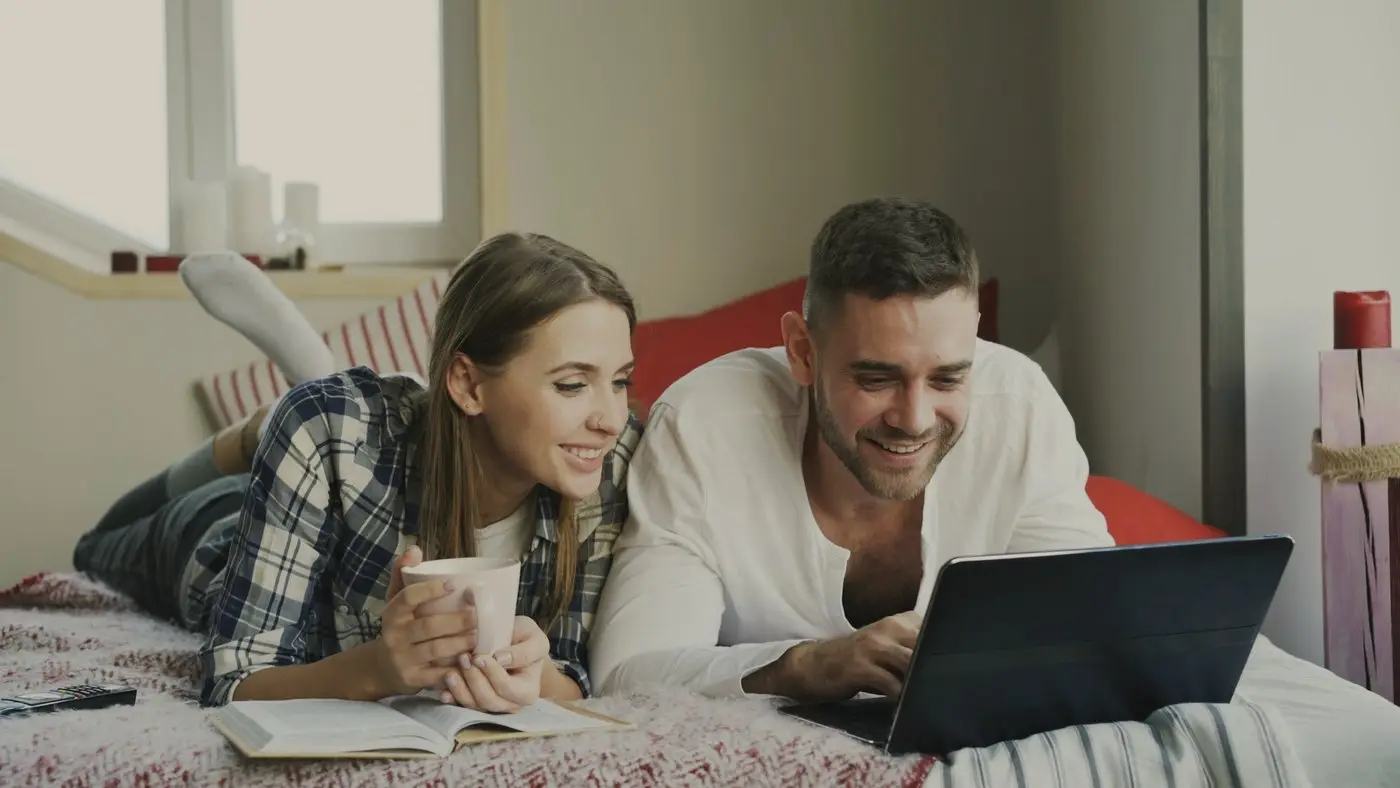 A couple lying on their bed with a laptop and coffee, smiling as they read through wedding photographers