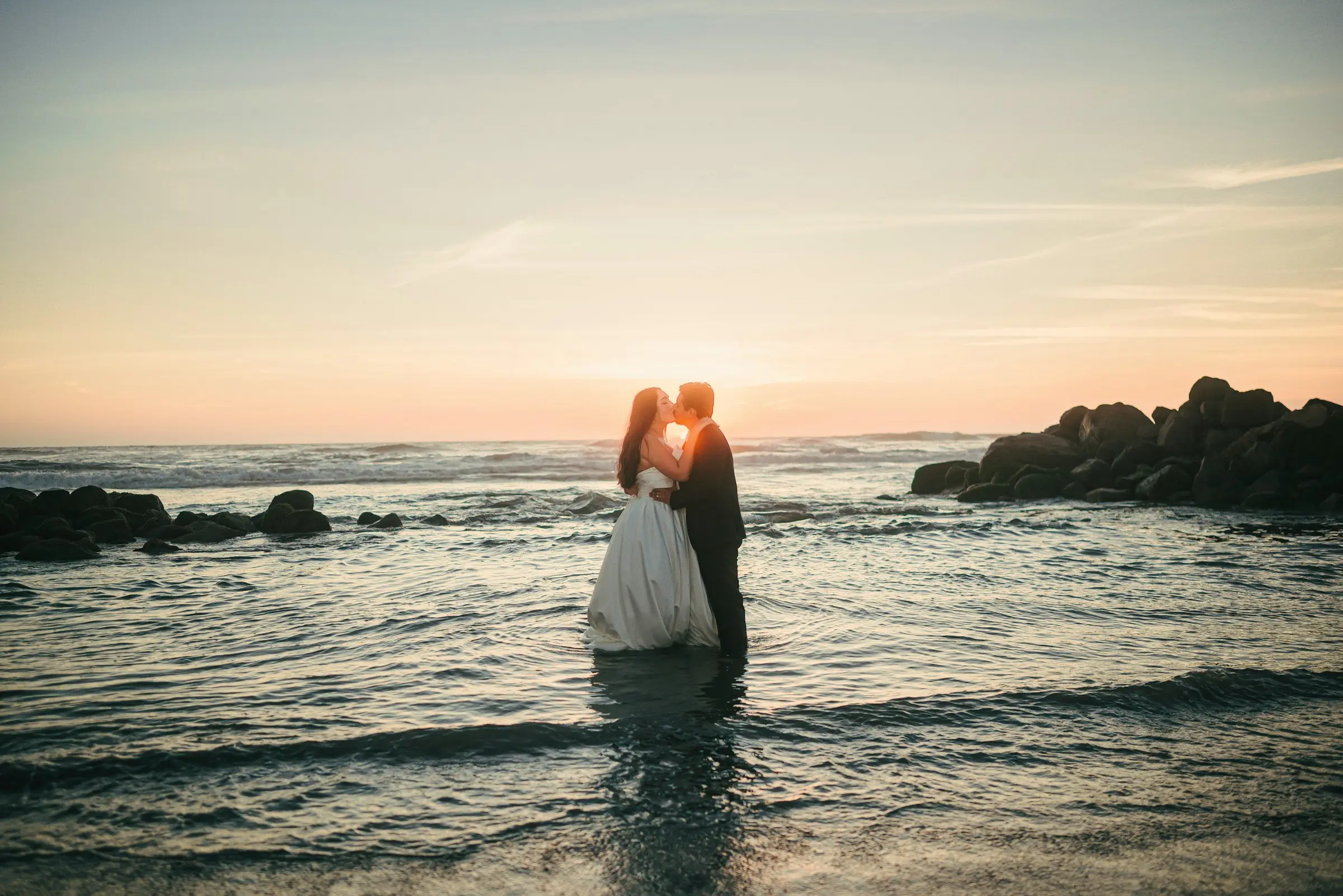 Bride and groom kiss standing ankle-deep in the surf, backlit by a low sun over the ocean, dark rocks on the horizon.