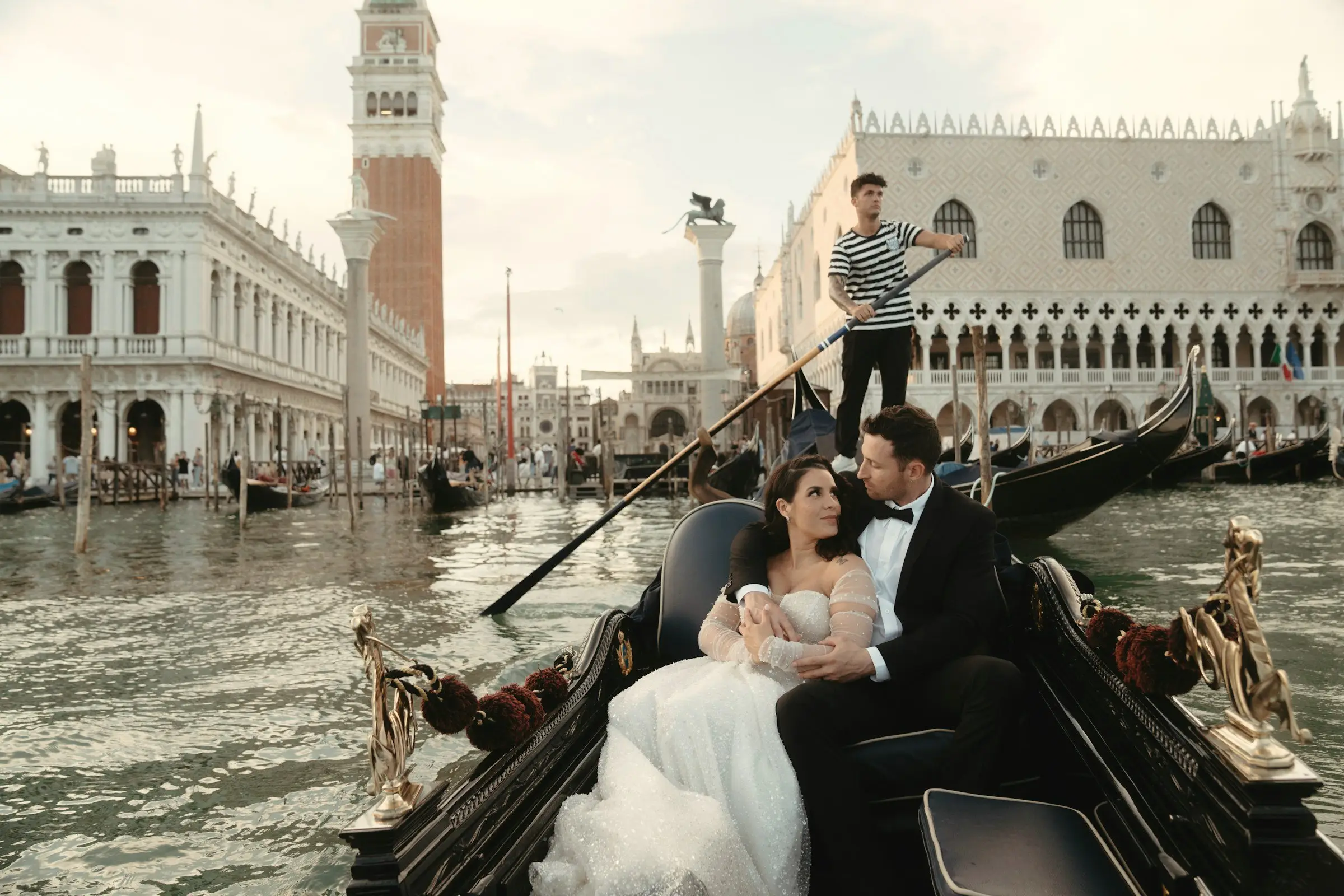 Bride and groom relax in a gondola on the water by St. Mark's Square, the campanile and Doge's Palace behind, gondolier standing at the stern.