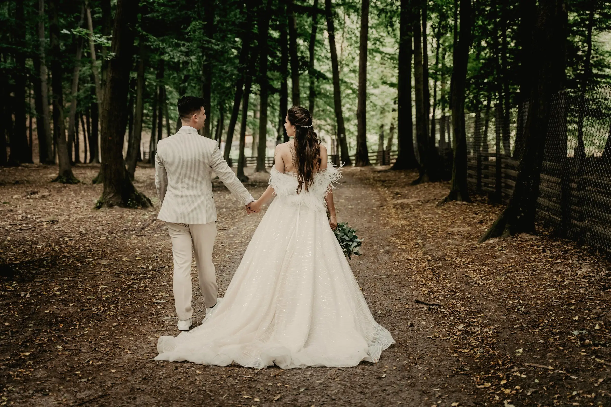 Couple walks hand in hand away from the camera down a shaded forest lane lined with tall pines.