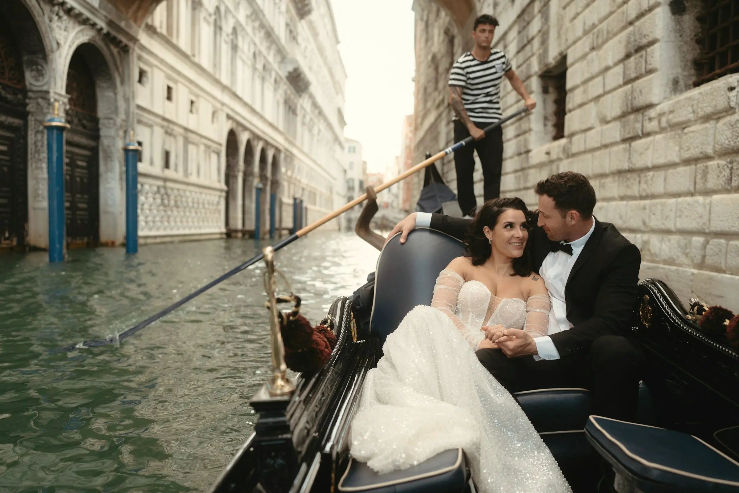 Couple smiles in a black gondola gliding down a narrow canal between pale stone palazzos, gondolier behind them.