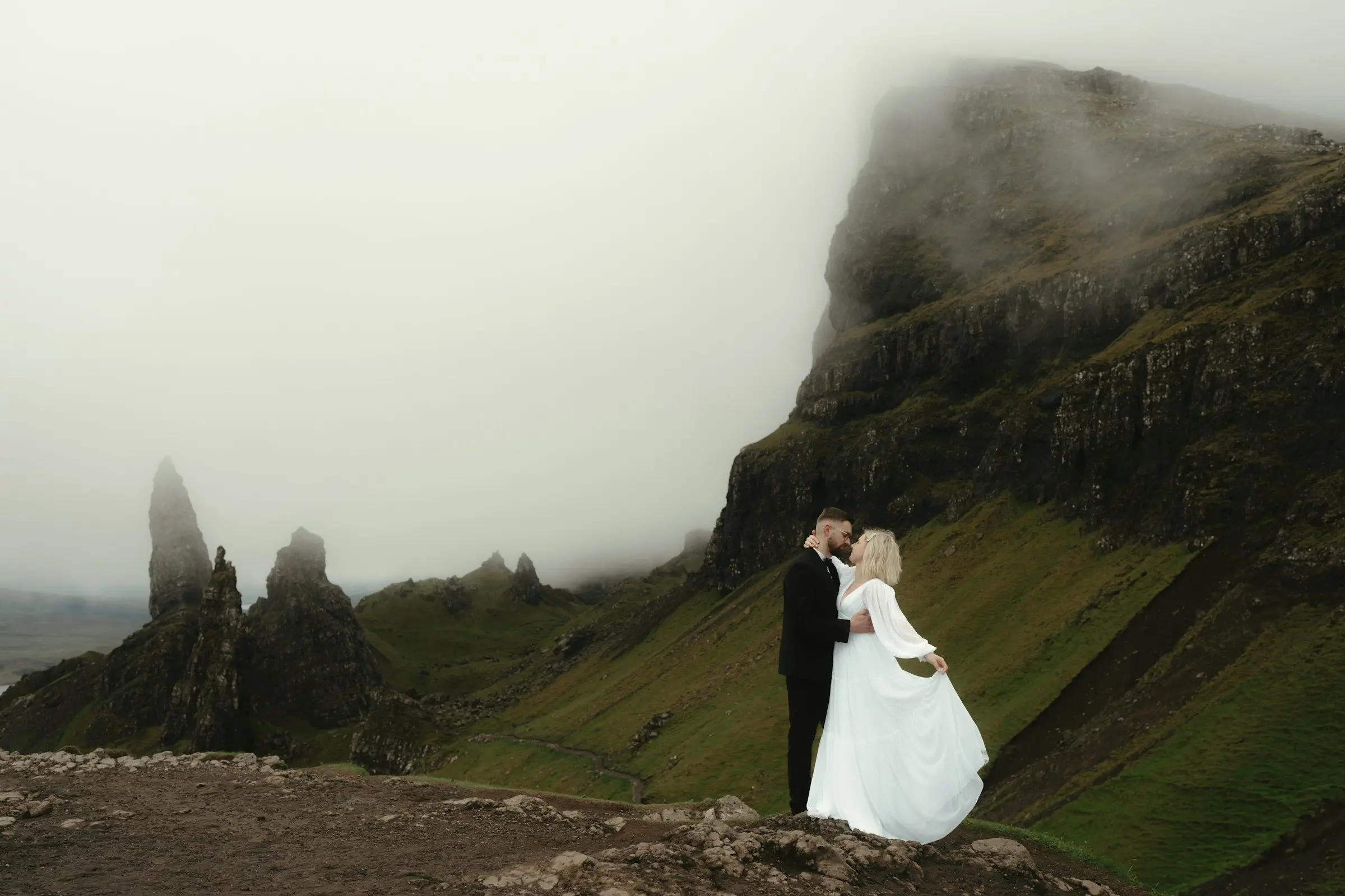 Couple kisses on a rocky outcrop beneath towering green cliffs disappearing into low cloud.