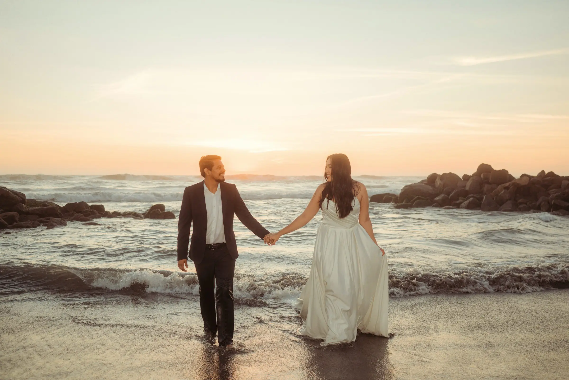 Bride and groom hold hands and wade out into gentle waves at sunset, seen from behind against a pastel sky.
