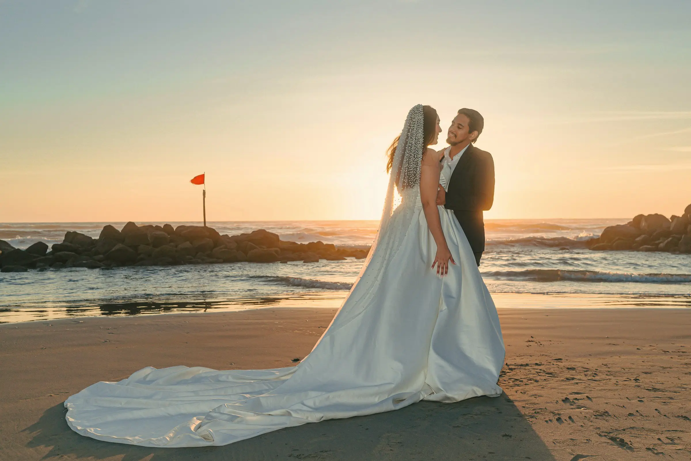 Bride with a long satin train spread across the sand stands with the groom by the water's edge, sun flaring low behind them.