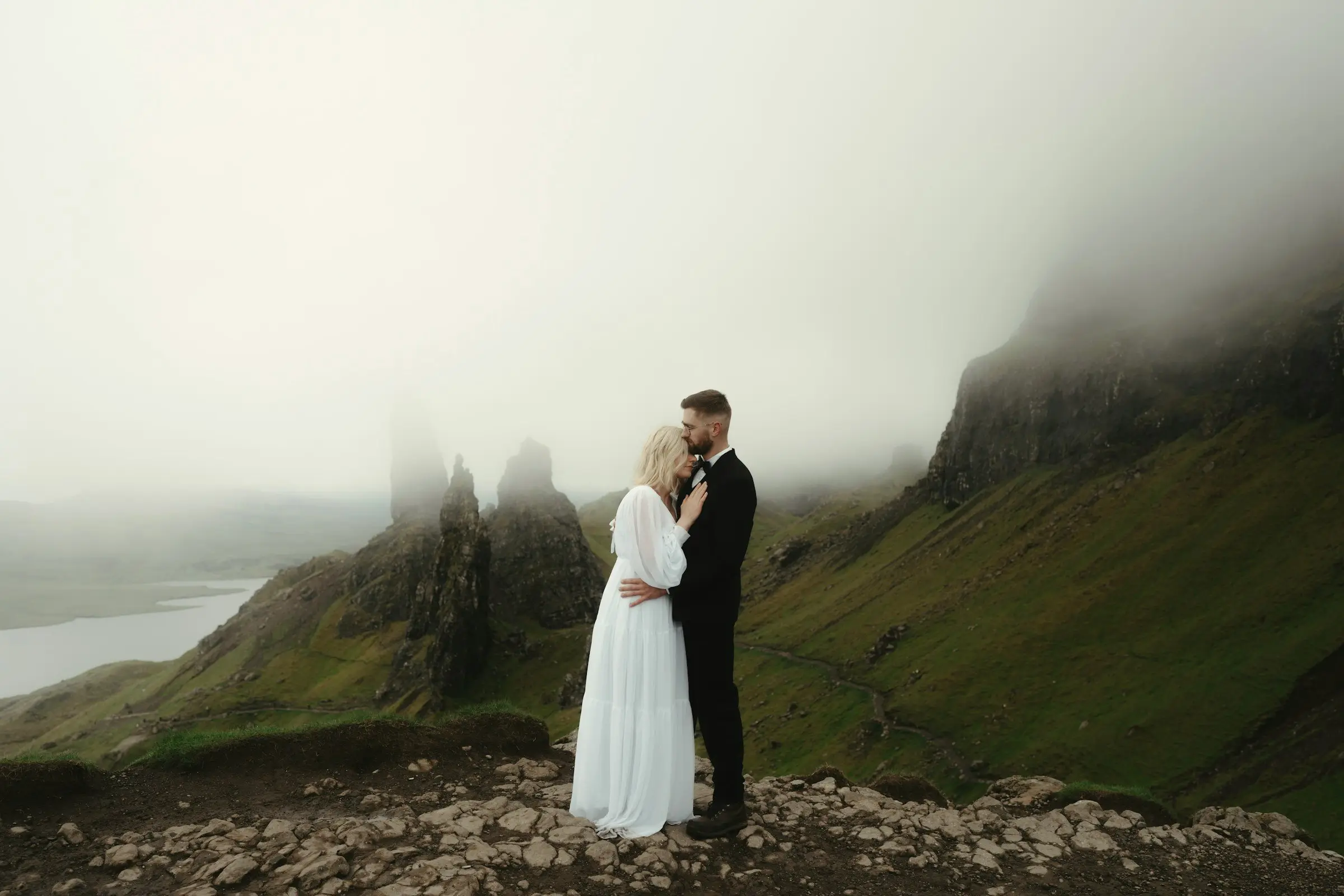 Bride and groom hold each other close on the trail with the jagged Storr pinnacles and a distant loch behind them in mist.