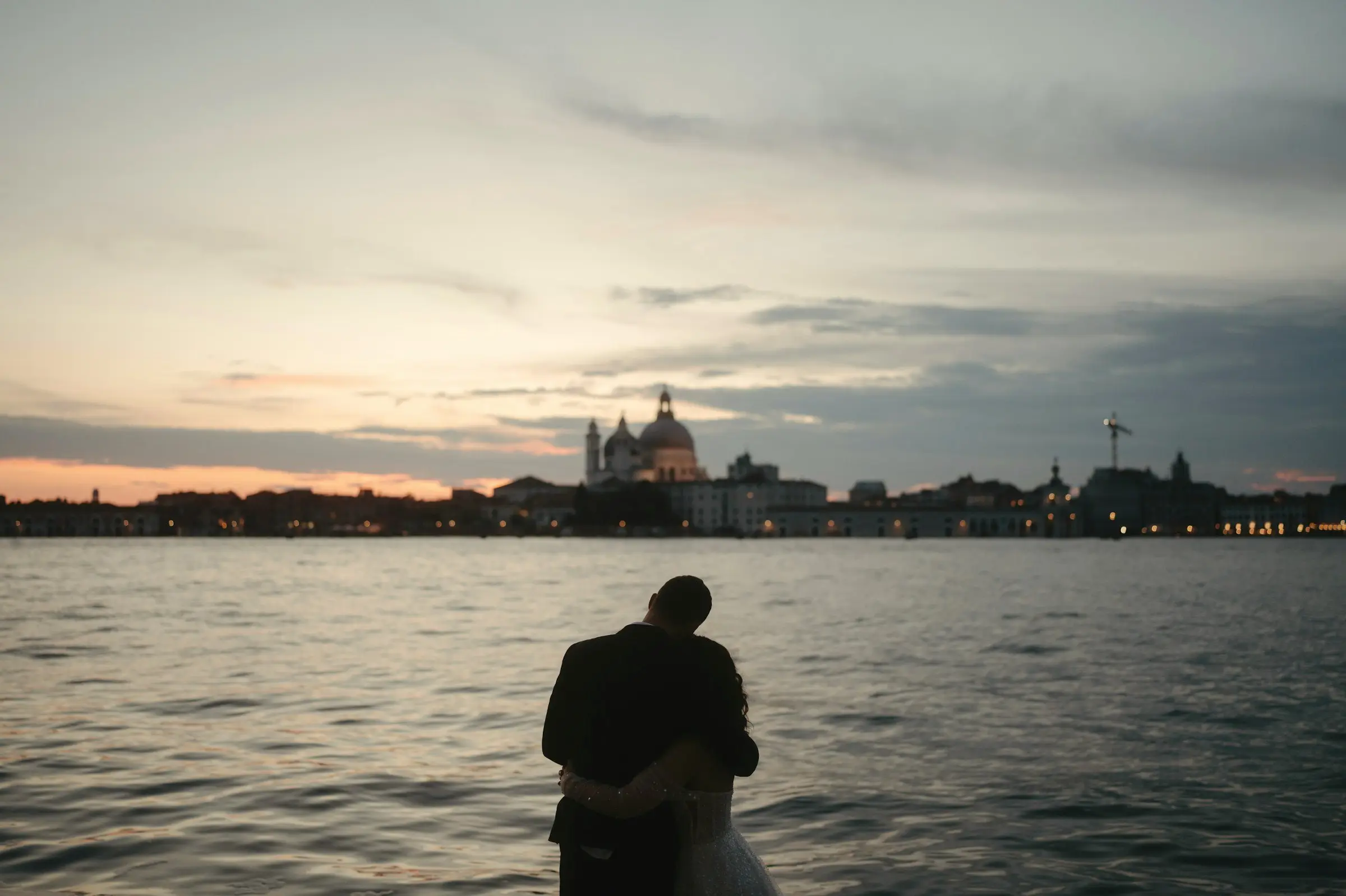 Small silhouette of the couple embracing at the water's edge with the Venice skyline and a pastel sunset sky beyond.