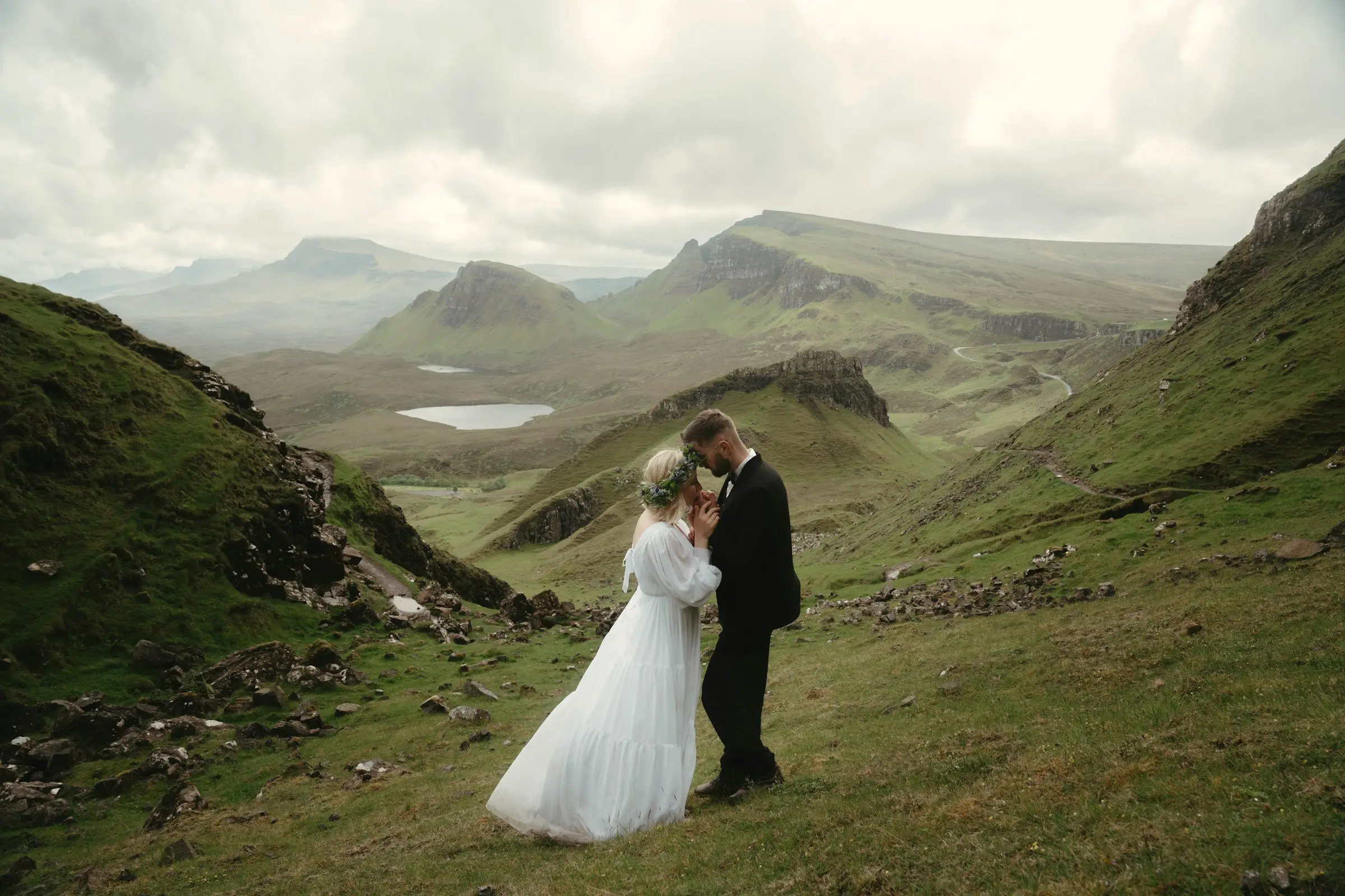 Couple embraces on a grassy slope overlooking the sweeping green valleys and small lochs of the Quiraing.
