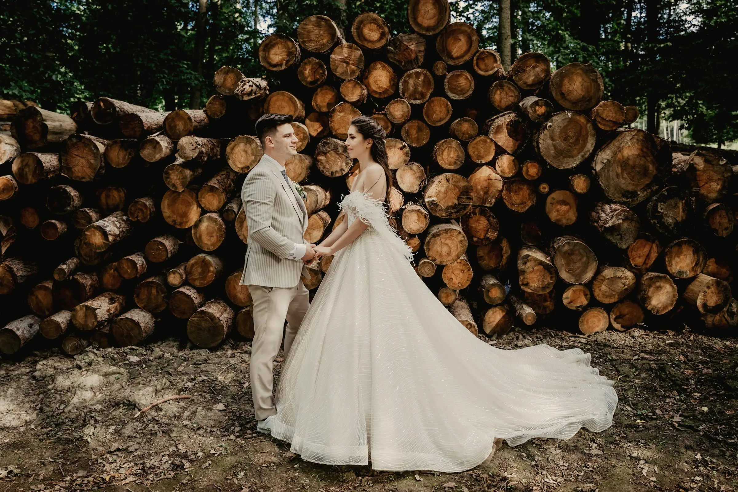 Bride and groom face each other in front of a tall stack of cut timber logs, her ball gown spread on the ground.