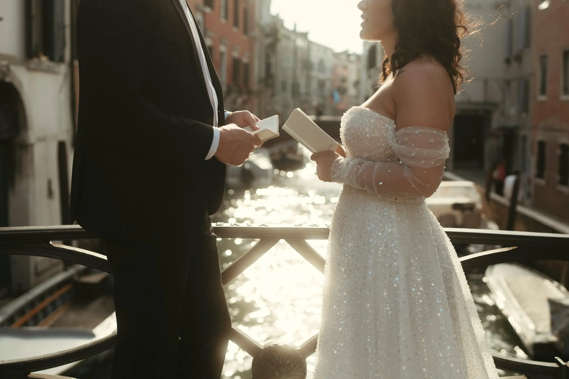 Bride in a sparkling gown holds her vow card facing the groom on a bridge over a sunlit Venetian canal.