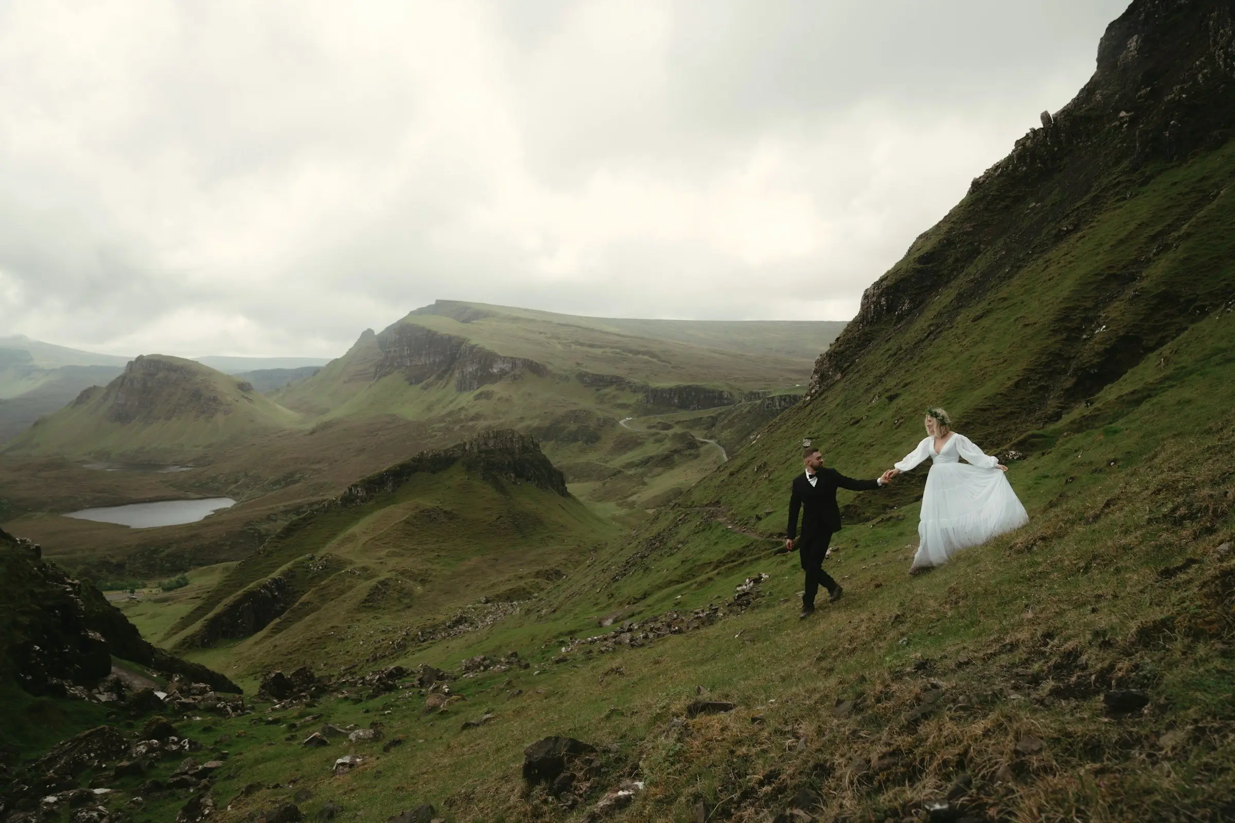 Groom and bride walk hand in hand up a steep green hillside, tiny against the vast Quiraing landscape.