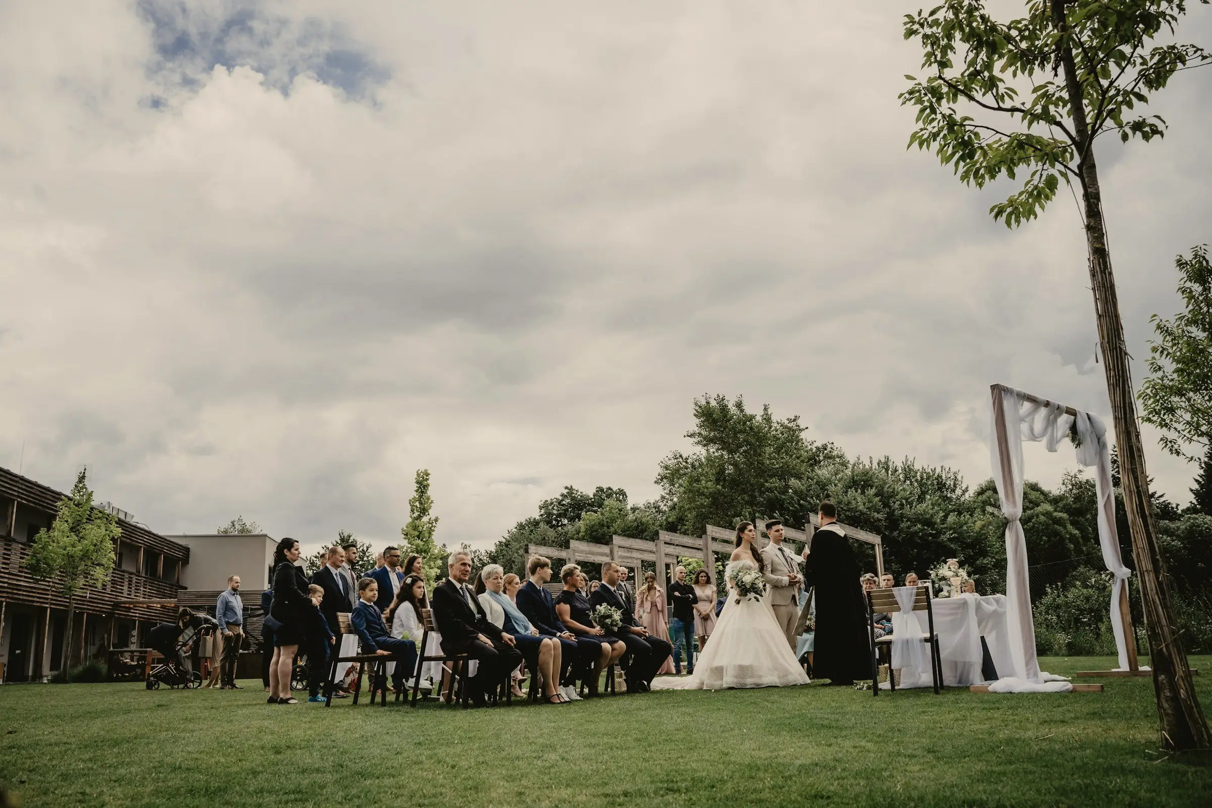 Wide view of an outdoor lawn ceremony with seated guests facing a white arch under a cloudy summer sky