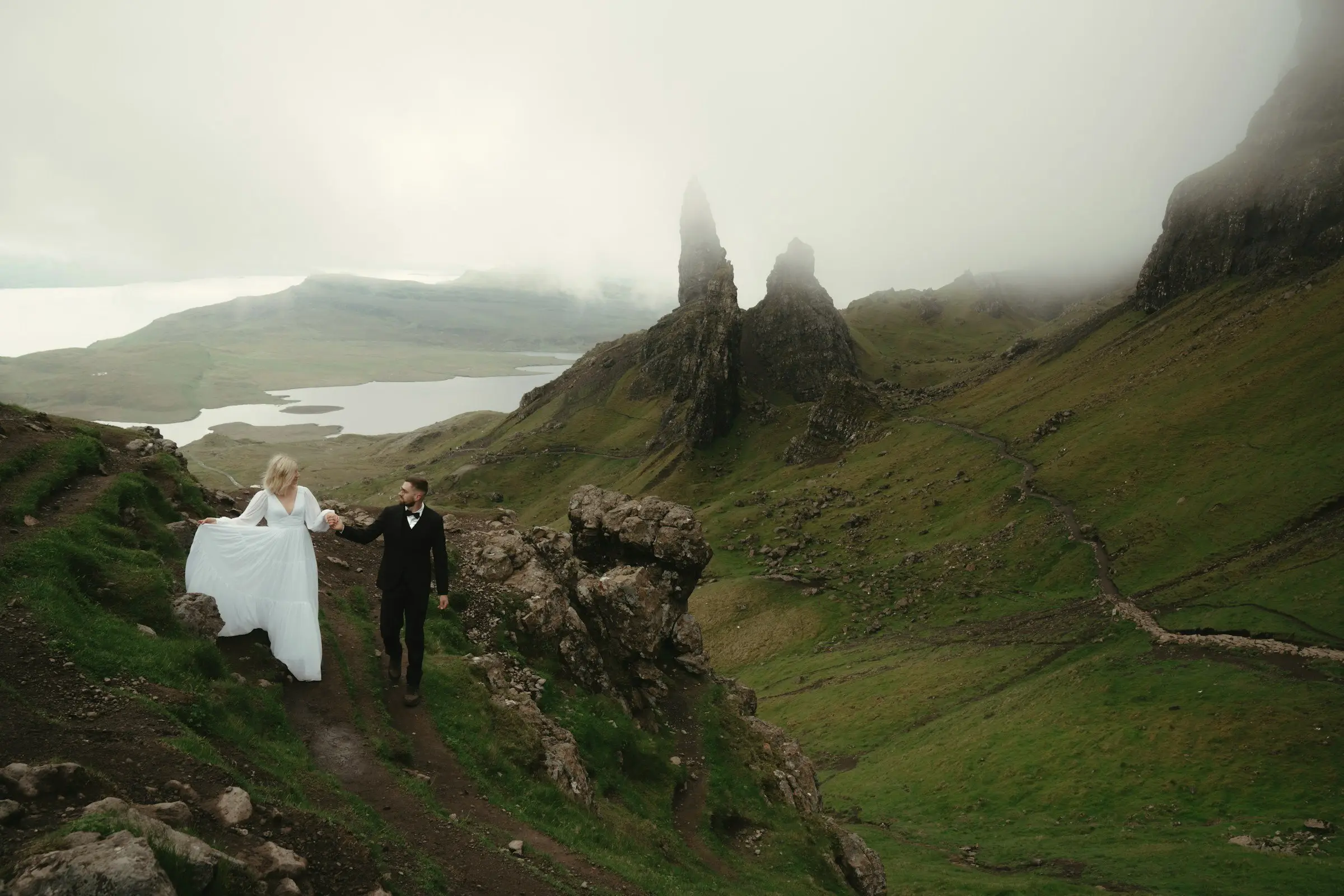 Bride lifts her dress as the couple walks along a winding path beneath the Storr rock spires.