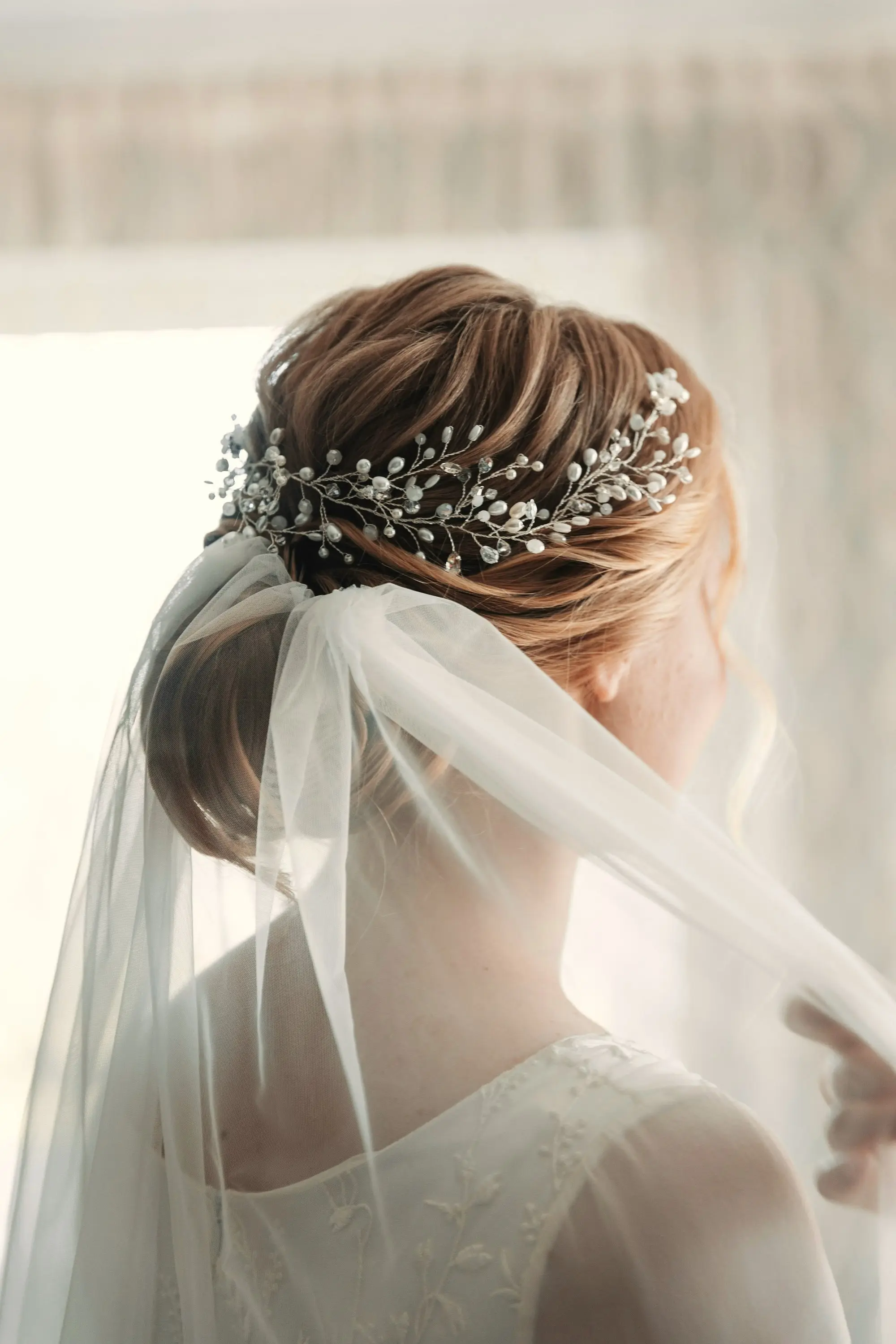 A bride seen from behind in soft window light, her veil and pearl hair vine glowing