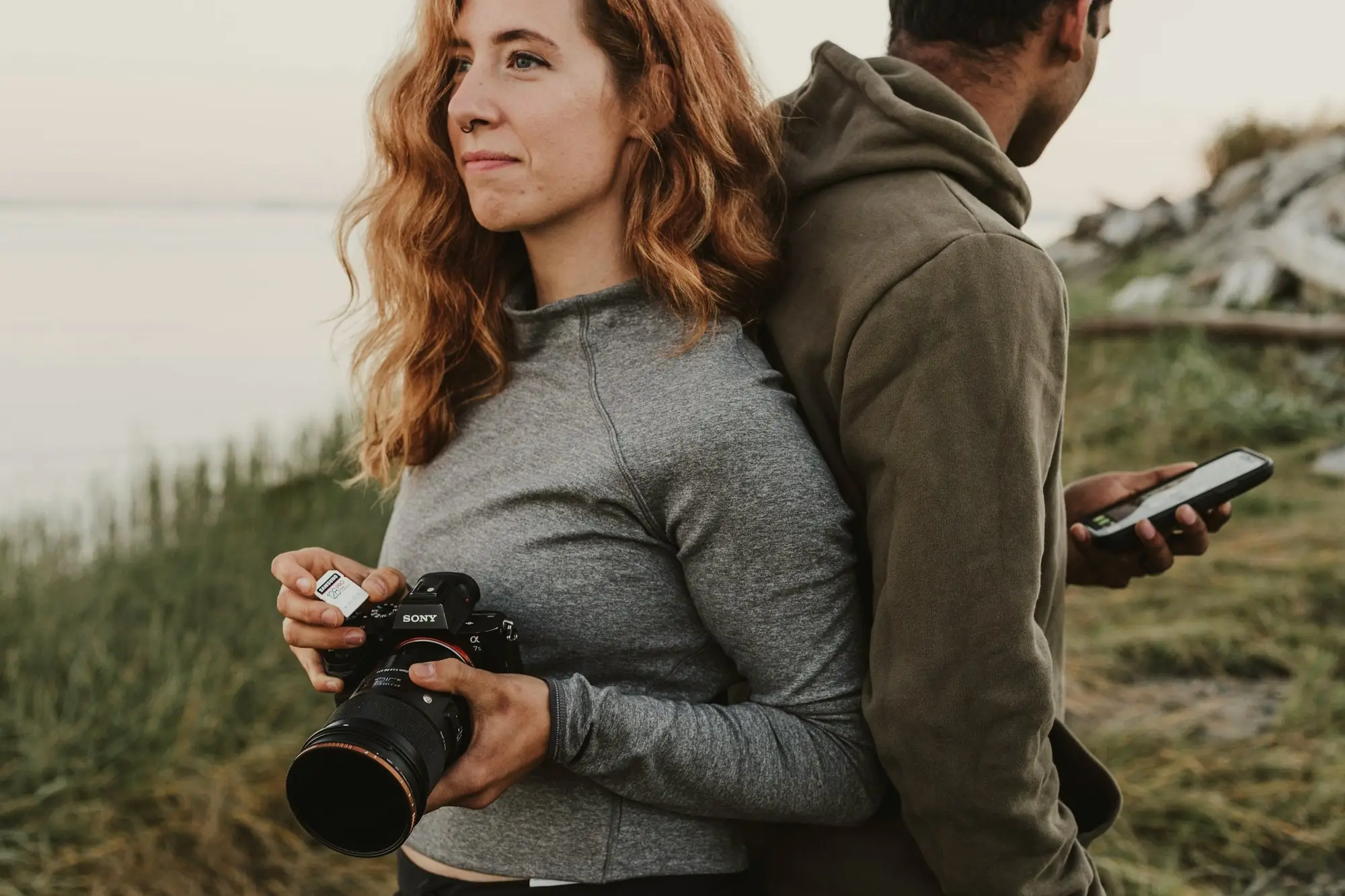 Claire, red-haired and holding her camera, leans back against Stephen on a grassy bluff above the sea
