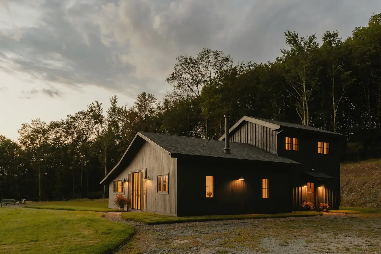 Modern two-story home with black and natural wood siding, warm lights on at dusk, a gravel drive and woods behind.