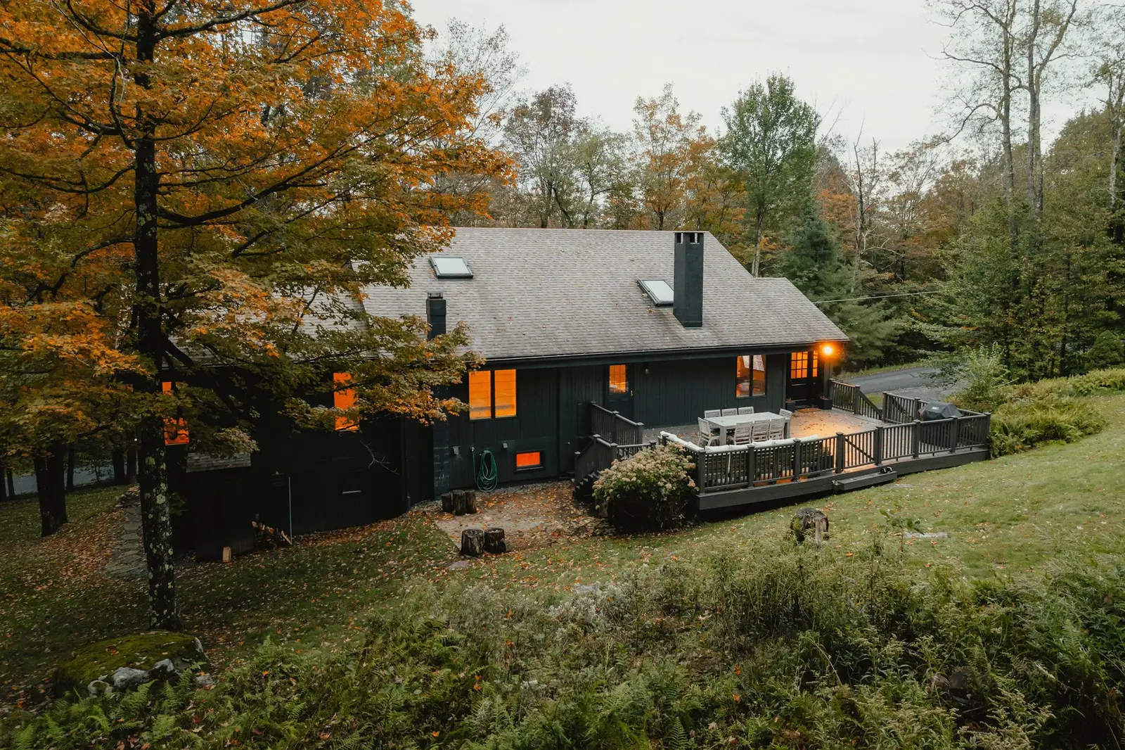 Dark green cabin with lit windows and a deck among autumn trees, a sloping lawn in front.
