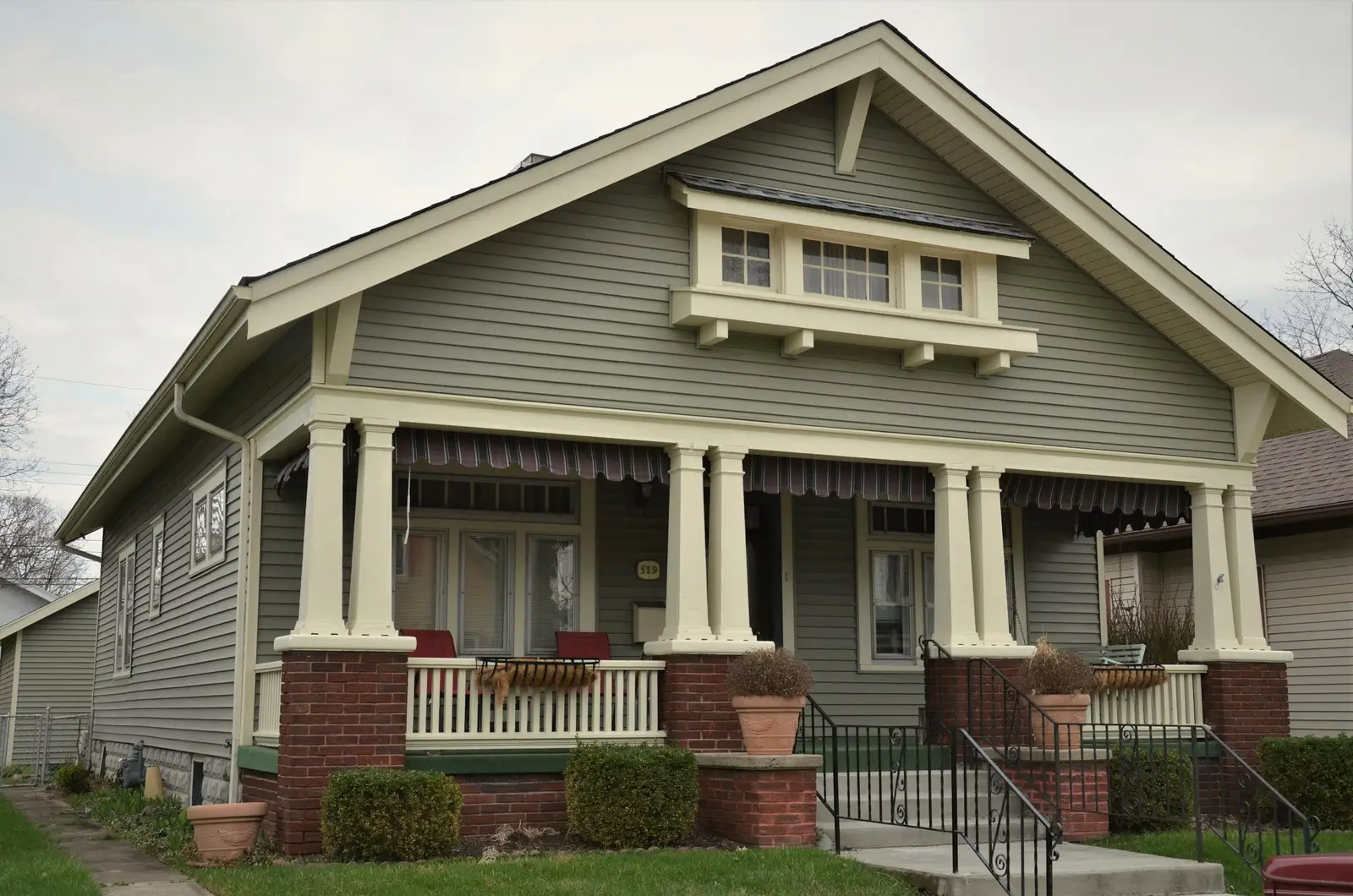 Gray-green Craftsman bungalow with tapered white columns on brick piers, a full-width front porch with striped awnings and potted plants on the steps.