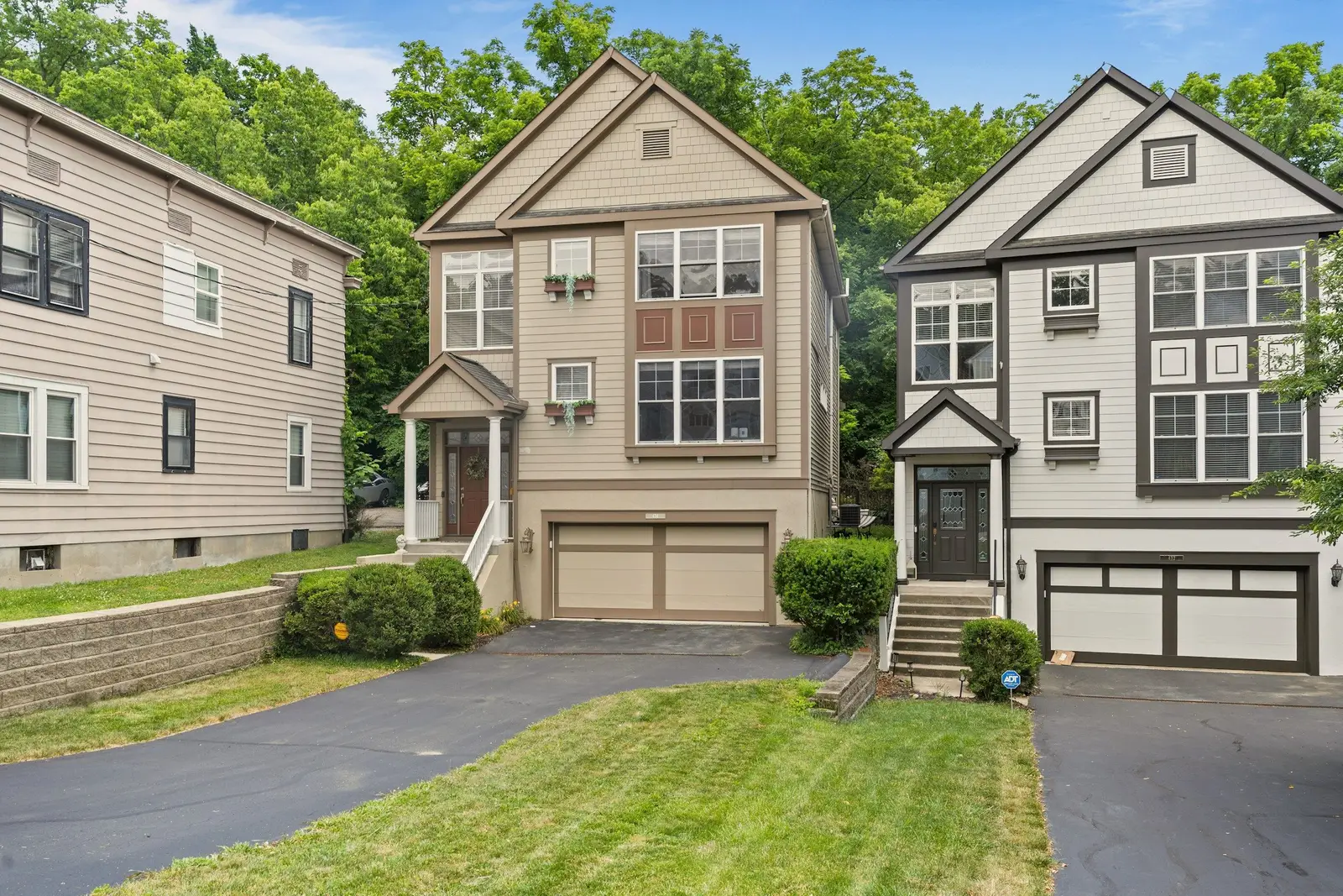 Two new three-story townhomes with beige and gray siding, two-car garages and a paved drive, trees behind.