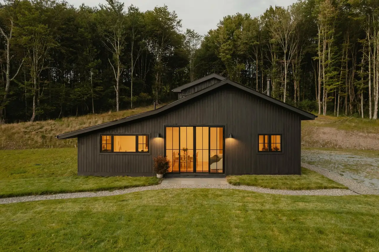 Dark-sided modern farmhouse with glowing windows and a black glass door on a wide lawn at dusk, woods behind.