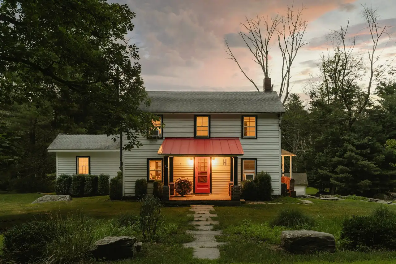 White two-story farmhouse at dusk with a red metal awning over a lit front door and a stone path across the lawn.