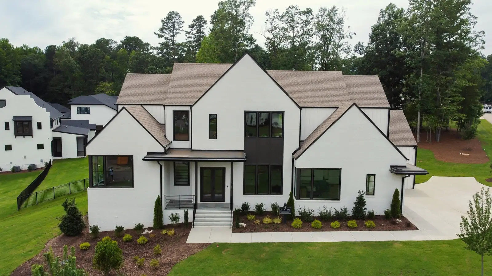 White modern farmhouse with steep gables, black-framed windows and a new lawn, woods and neighboring homes behind.