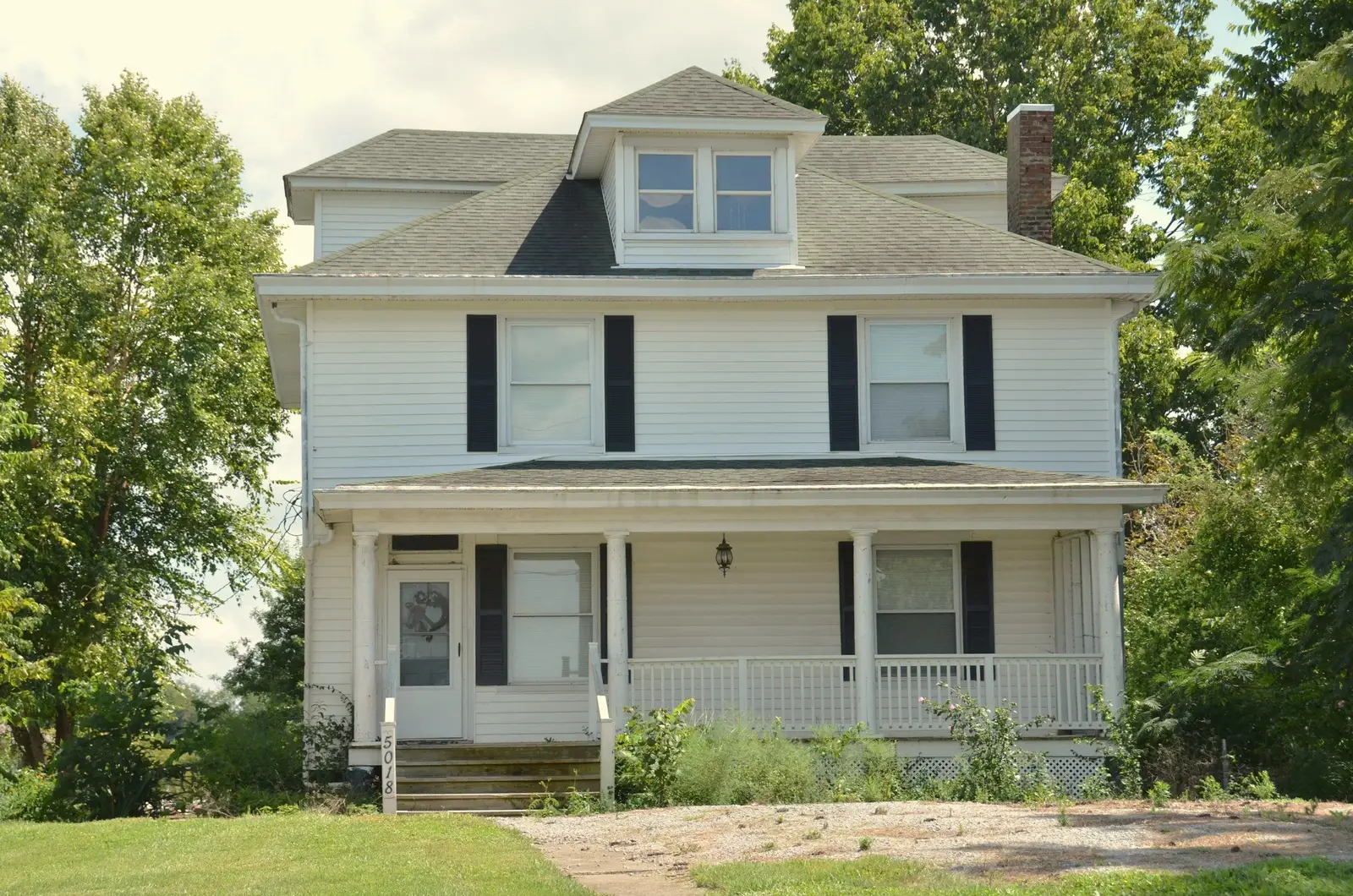 White Foursquare with black shutters, a hipped dormer and a full-width front porch, tall trees on both sides.