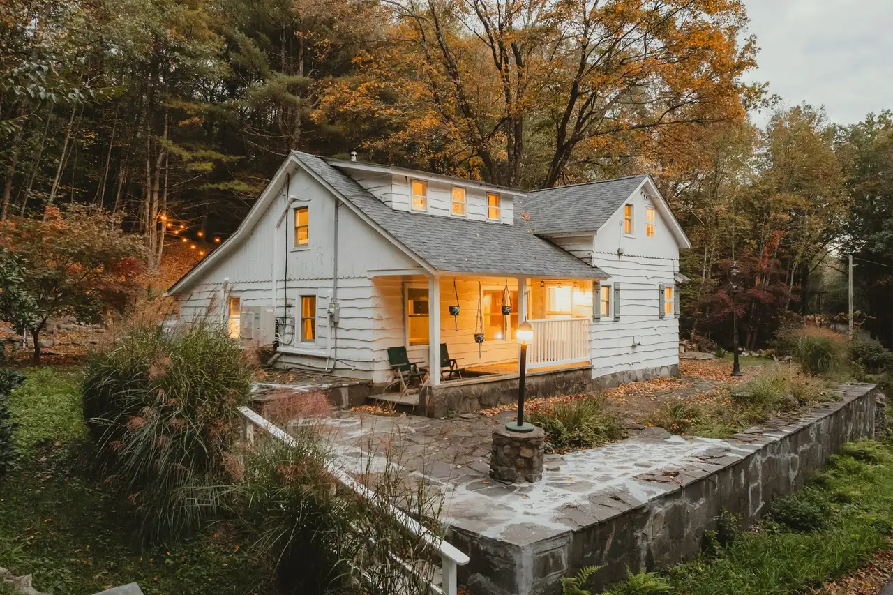 White cottage with lit windows and a covered porch at dusk, a stone retaining wall and autumn trees around it.