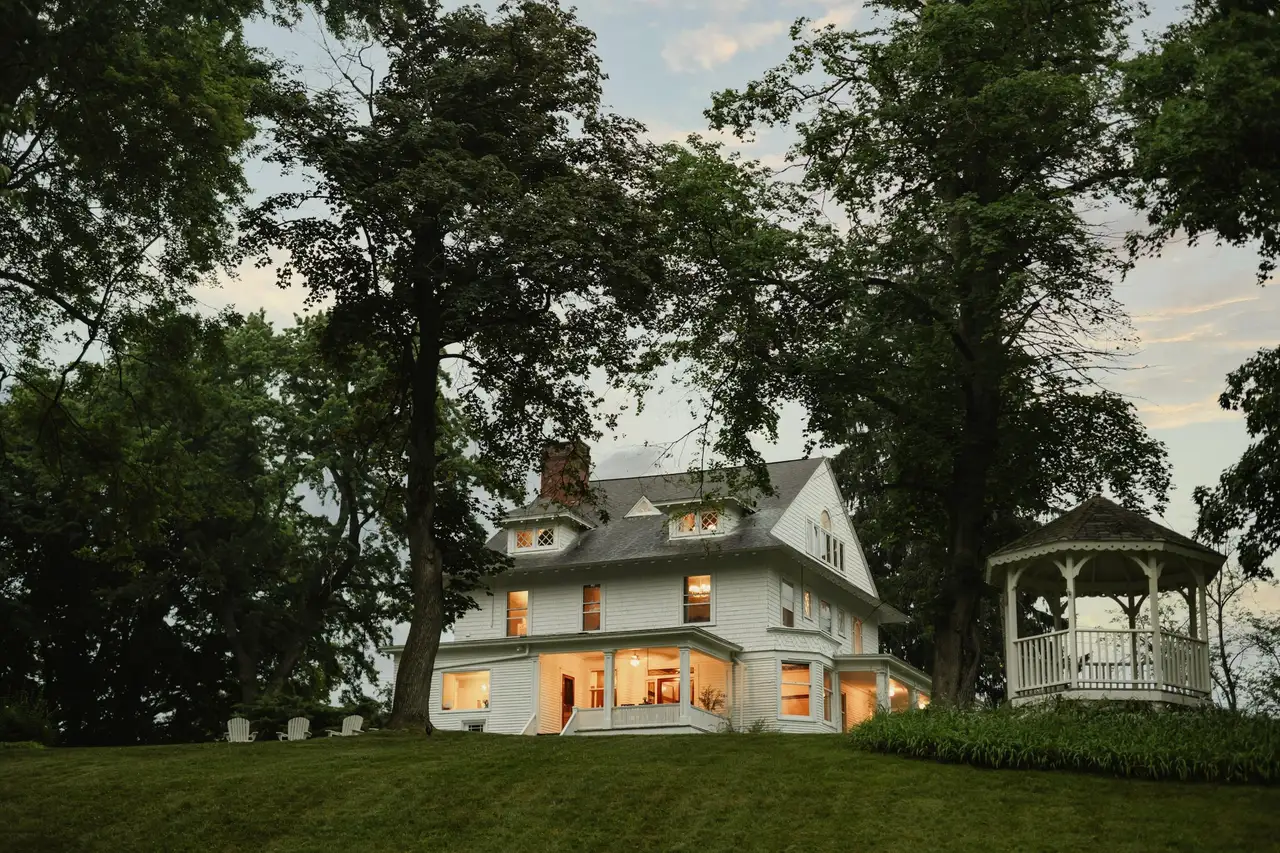 Large white Victorian with glowing windows at dusk, a wraparound porch, tall oaks and a white gazebo on the lawn.