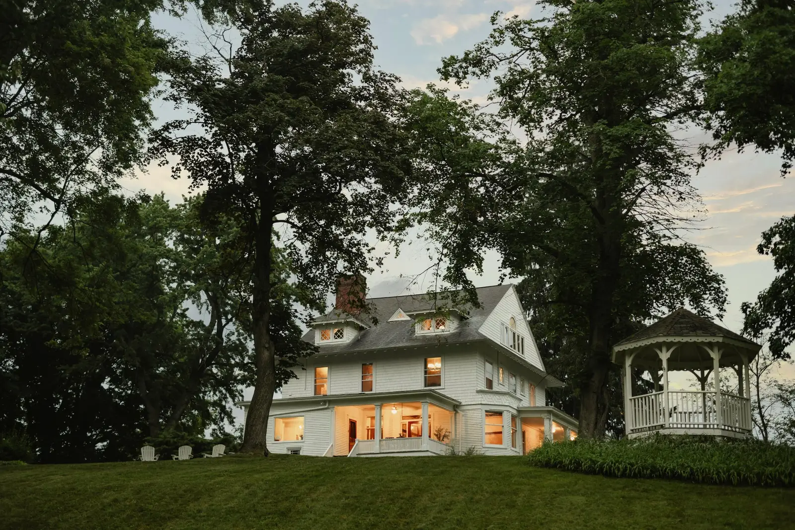 Large white Victorian with glowing windows at dusk, a wraparound porch, tall oaks and a white gazebo on the lawn.