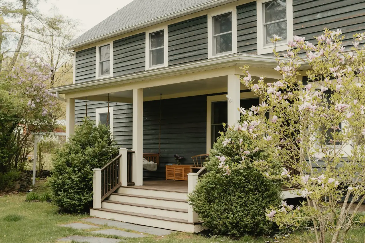 Two-story gray-blue Colonial with white trim, a deep covered front porch and wide steps, flowering shrubs in front.