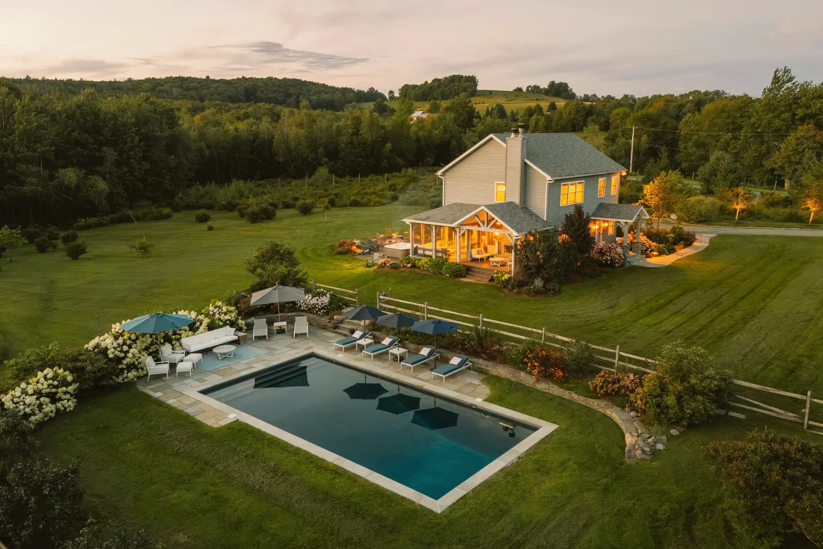 Aerial view at dusk of a farmhouse with a lit porch, a pool and a fenced lawn, wooded hills behind.