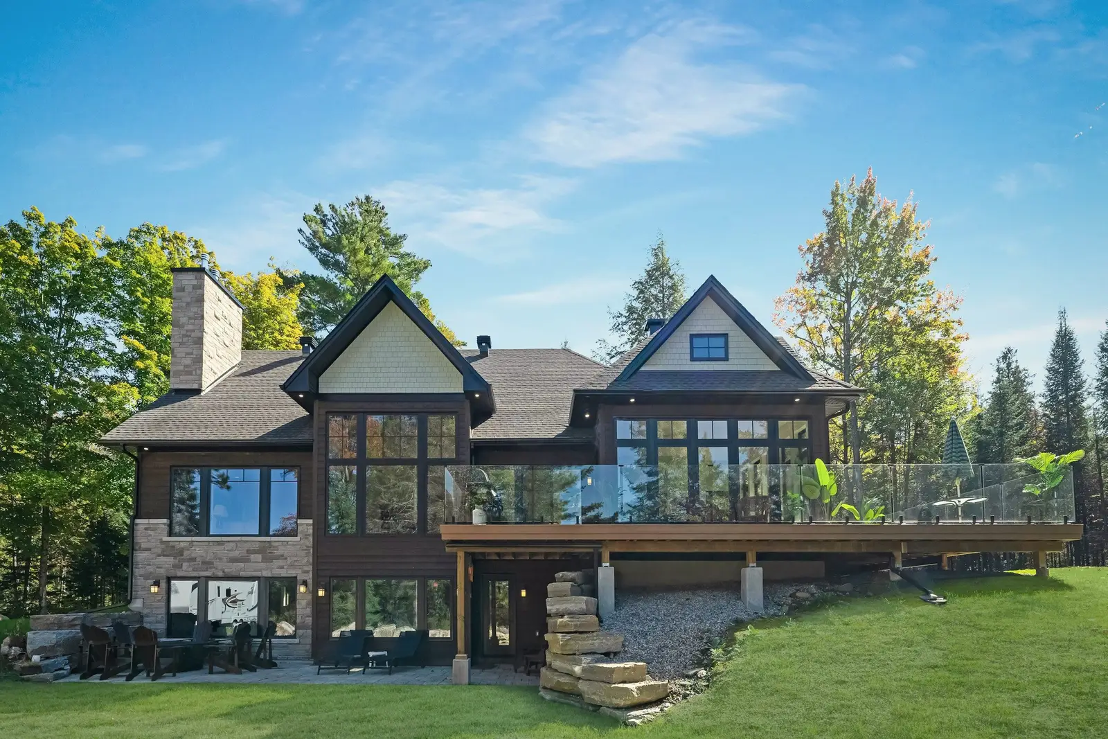 Two-story lake home with stone and dark wood siding, tall glass gables and a long deck with glass railings above a green lawn.
