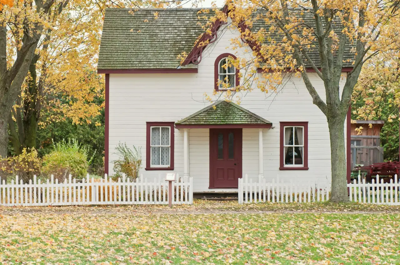 White cottage with a dark red front door, an arched upstairs window and a white picket fence, autumn leaves on the lawn.