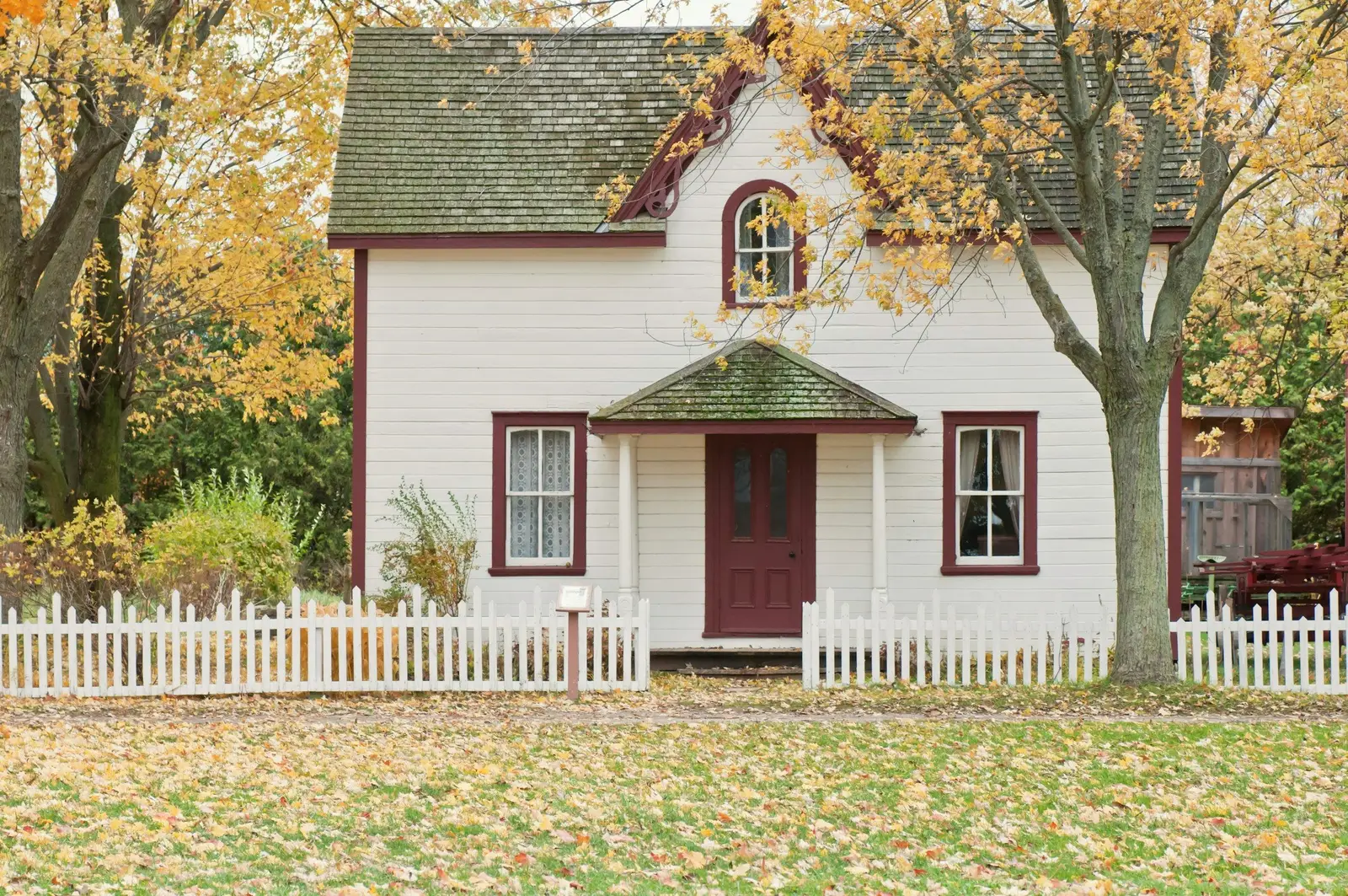 White cottage with a dark red front door, an arched upstairs window and a white picket fence, autumn leaves on the lawn.