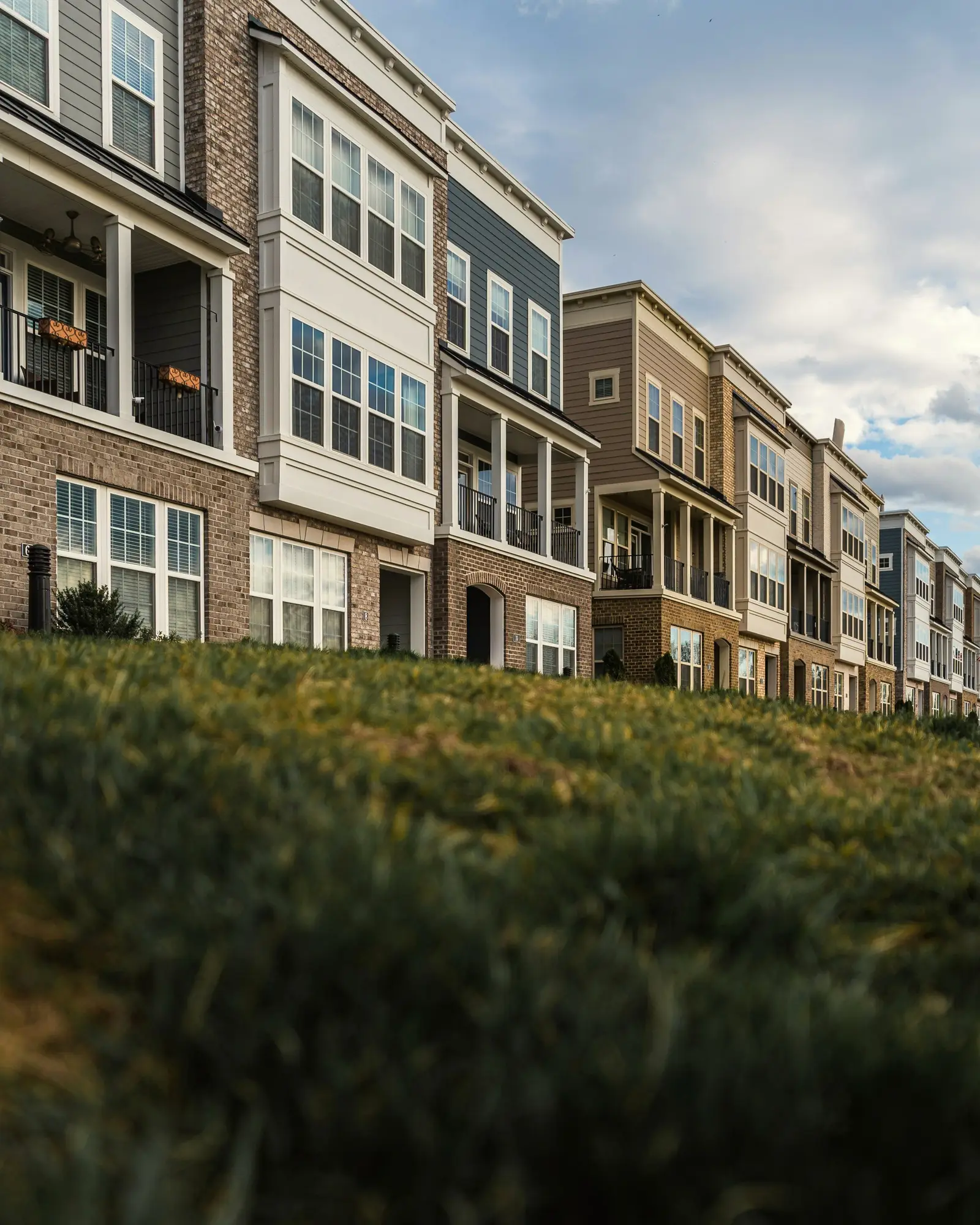 Row of three-story townhomes with brick lower floors, light siding and covered balconies above a grassy slope.