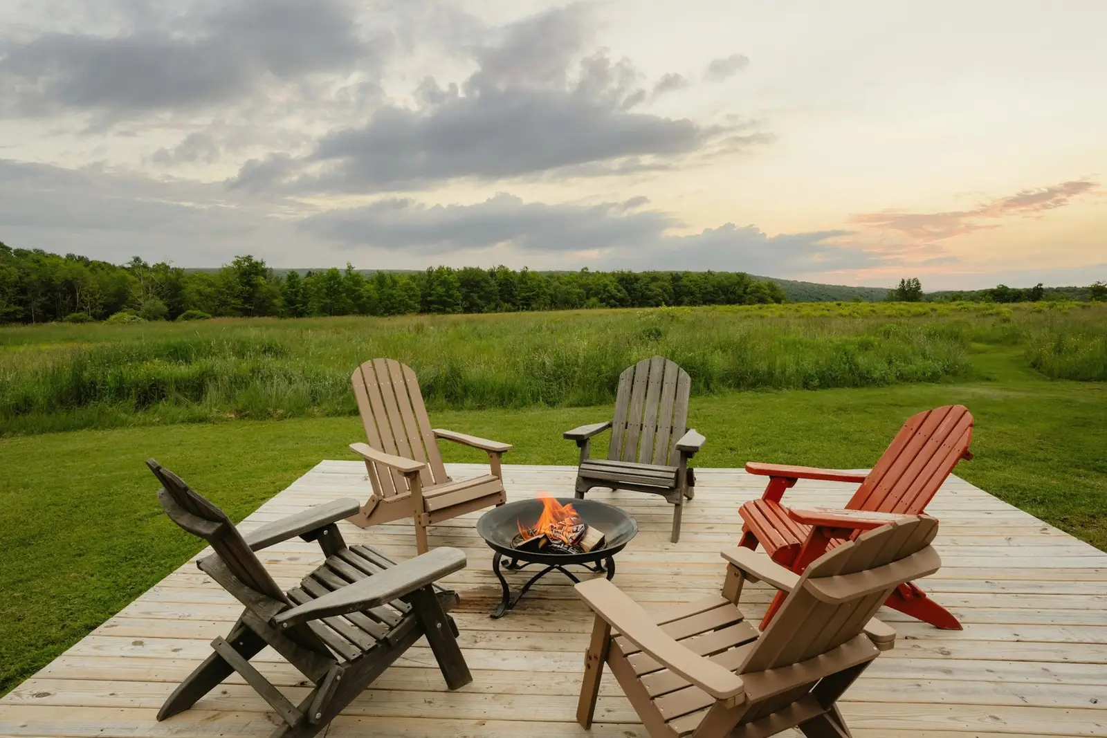 Wooden terrace with Adirondack chairs around a fire pit, a green meadow beyond.