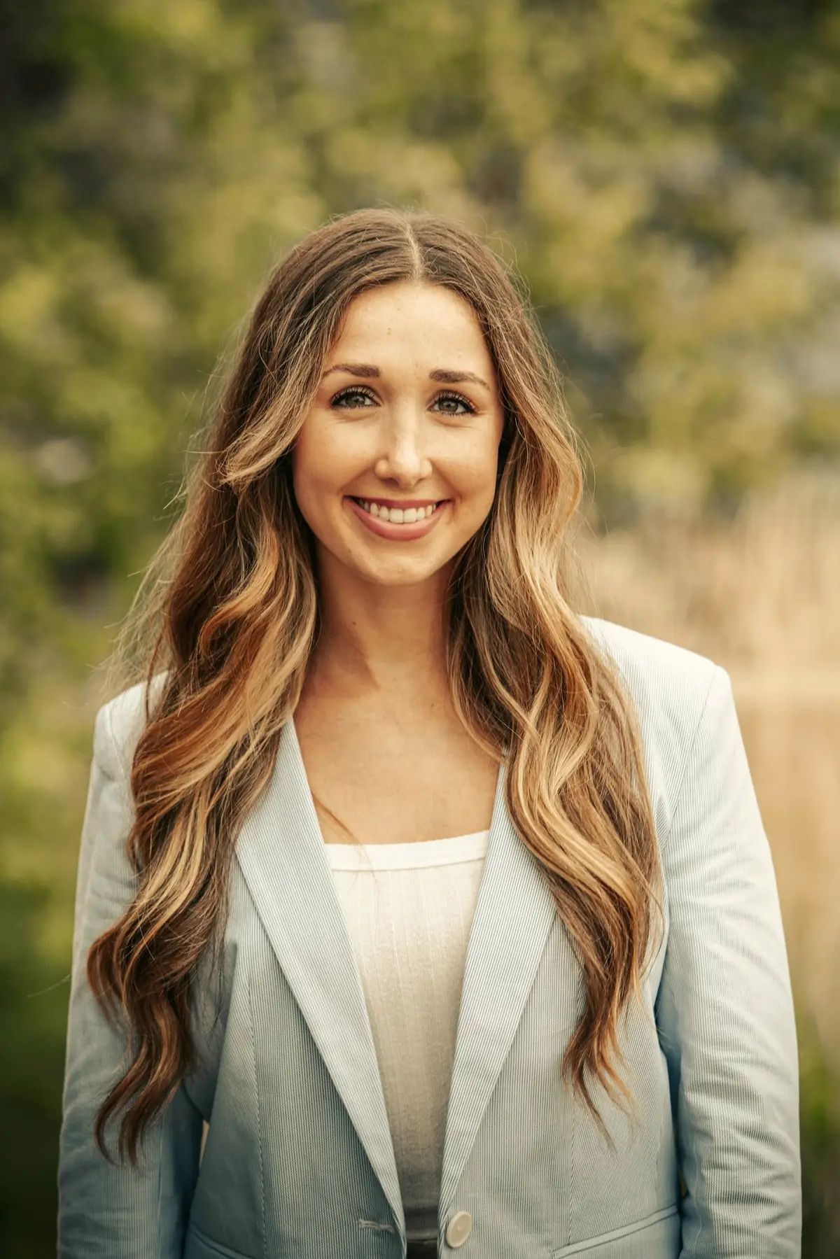 Portrait of Claire Bennett, smiling, with long brown hair and a light blazer, trees softly blurred behind her.