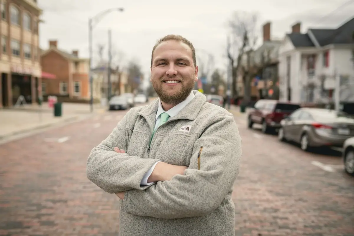Portrait of David Hart, smiling with arms crossed, standing on a brick main street lined with shops.