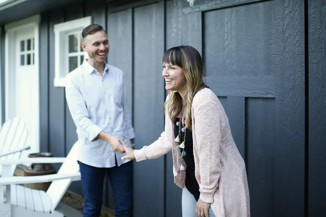 A Realtor shaking hands with a smiling client outside the front door of a dark-sided house.