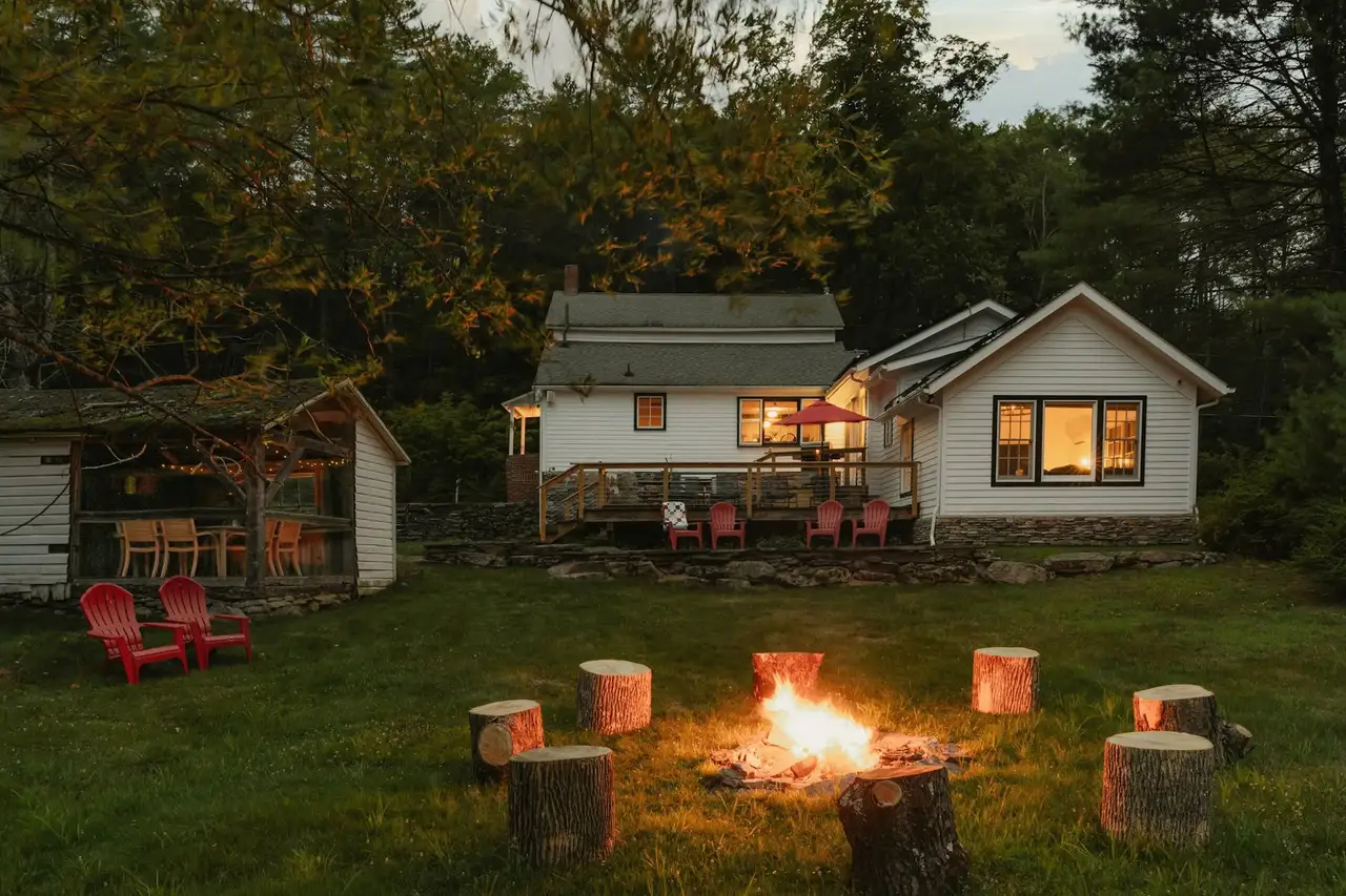 A white cottage with lit windows at dusk, red Adirondack chairs and a crackling fire pit on the lawn.