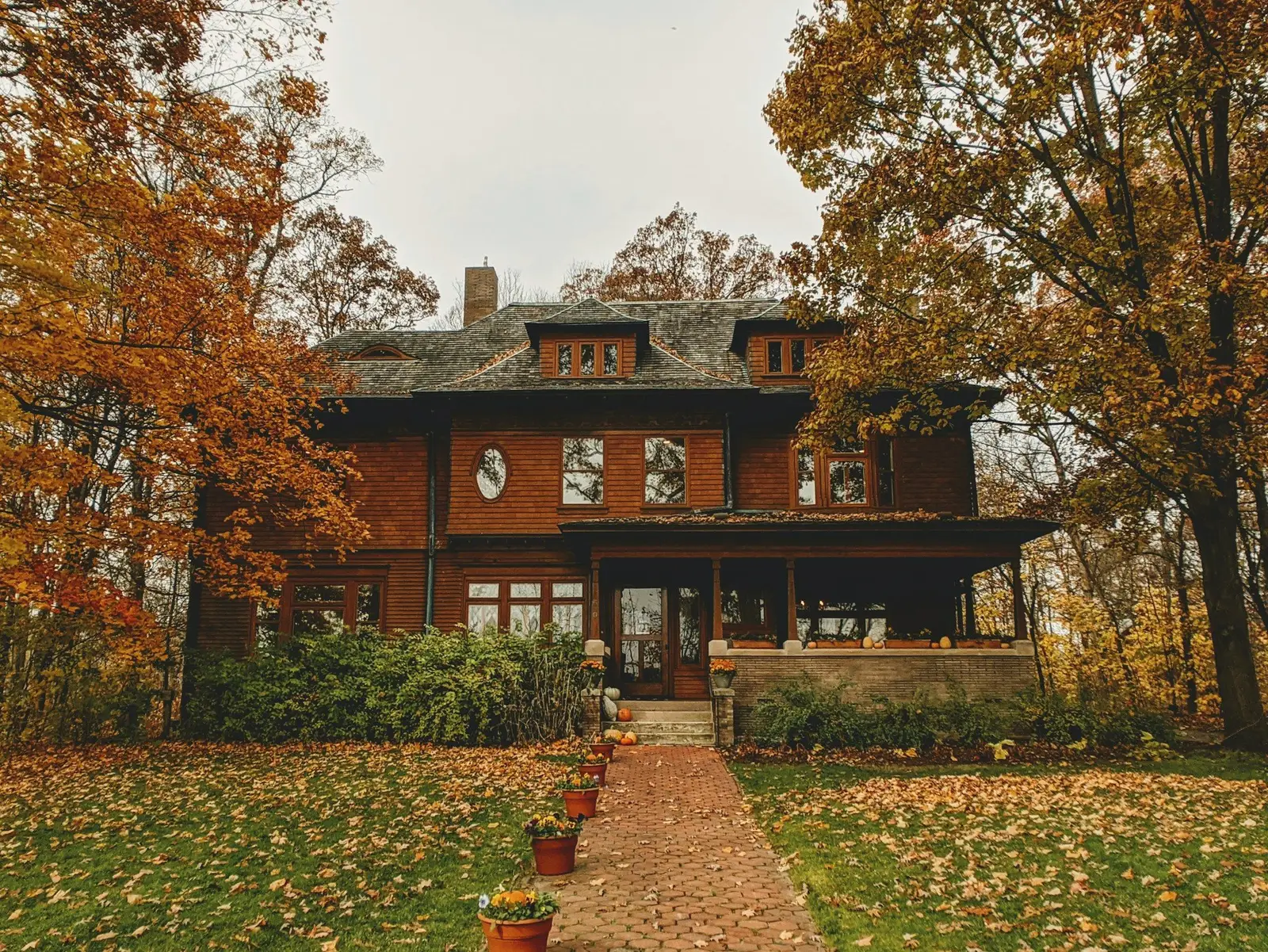 A brick house with a deep porch behind a lawn covered in autumn leaves, orange and gold trees around it.